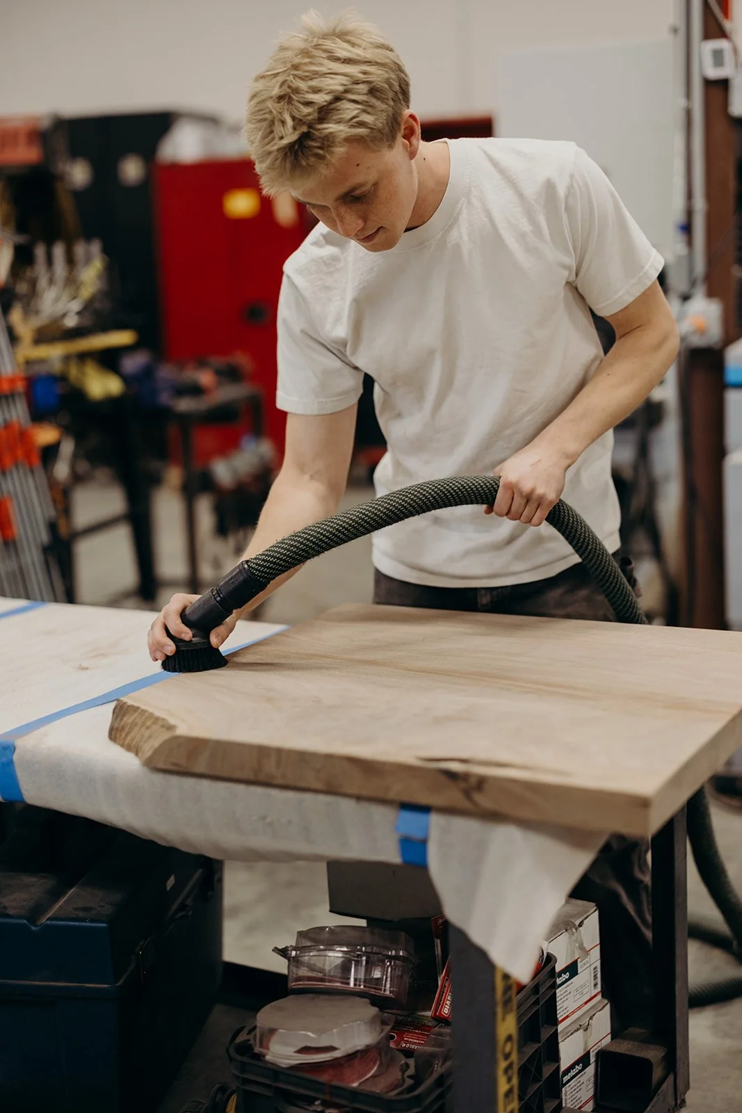 Young man using a vacuum cleaner on a large wooden piece in a workshop.