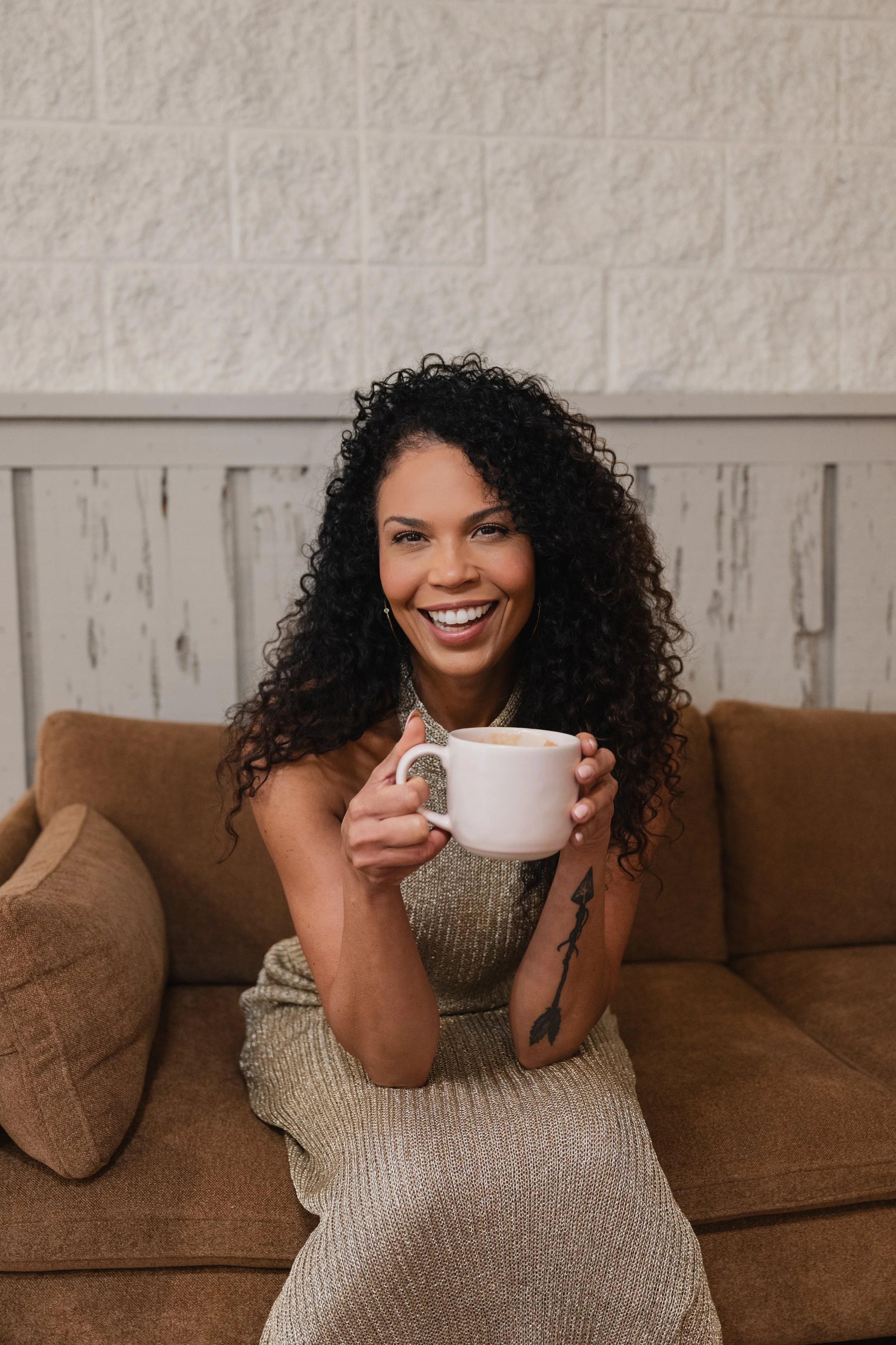 A woman with curly dark hair, smiling and holding a white mug, sitting on a brown couch in a cozy indoor setting.