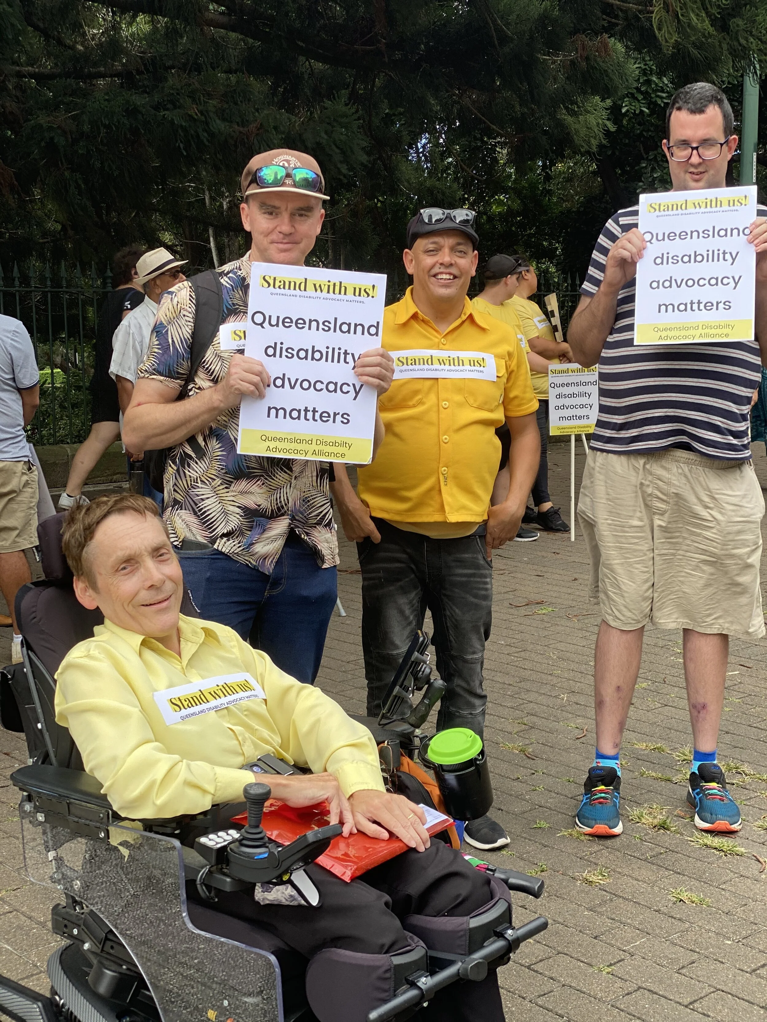 Man sitting in wheelchair in yellow shirt and three men standing beside him,  two holding signs large sign with the Stand with us! logo at the top and it says Queensland disability advocacy matters. 