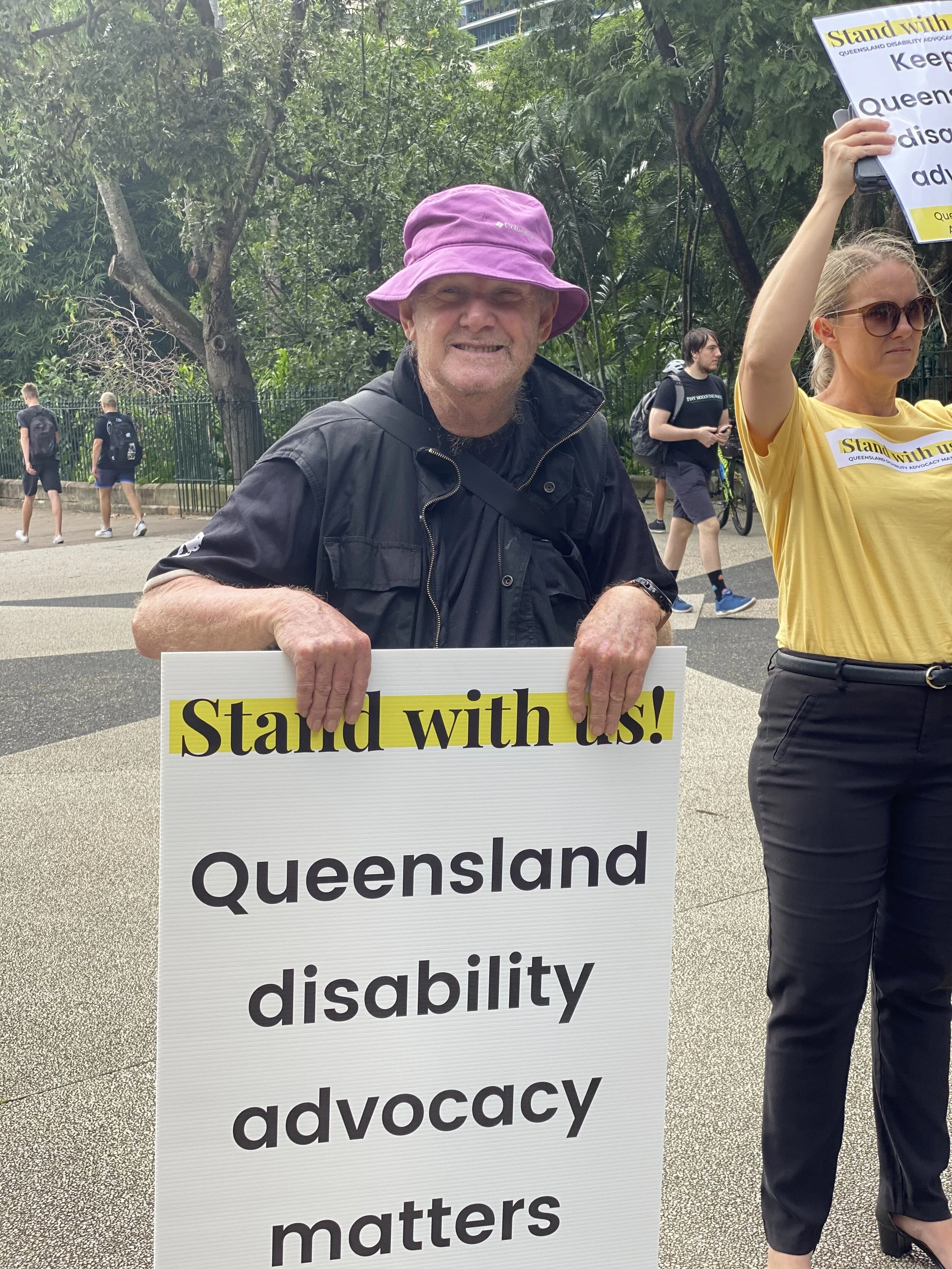 A man holding a large sign with the Stand with us! logo at the top and it says Queensland disability advocacy matters. There is a woman in a yellow shirt standing beside holding sign 
