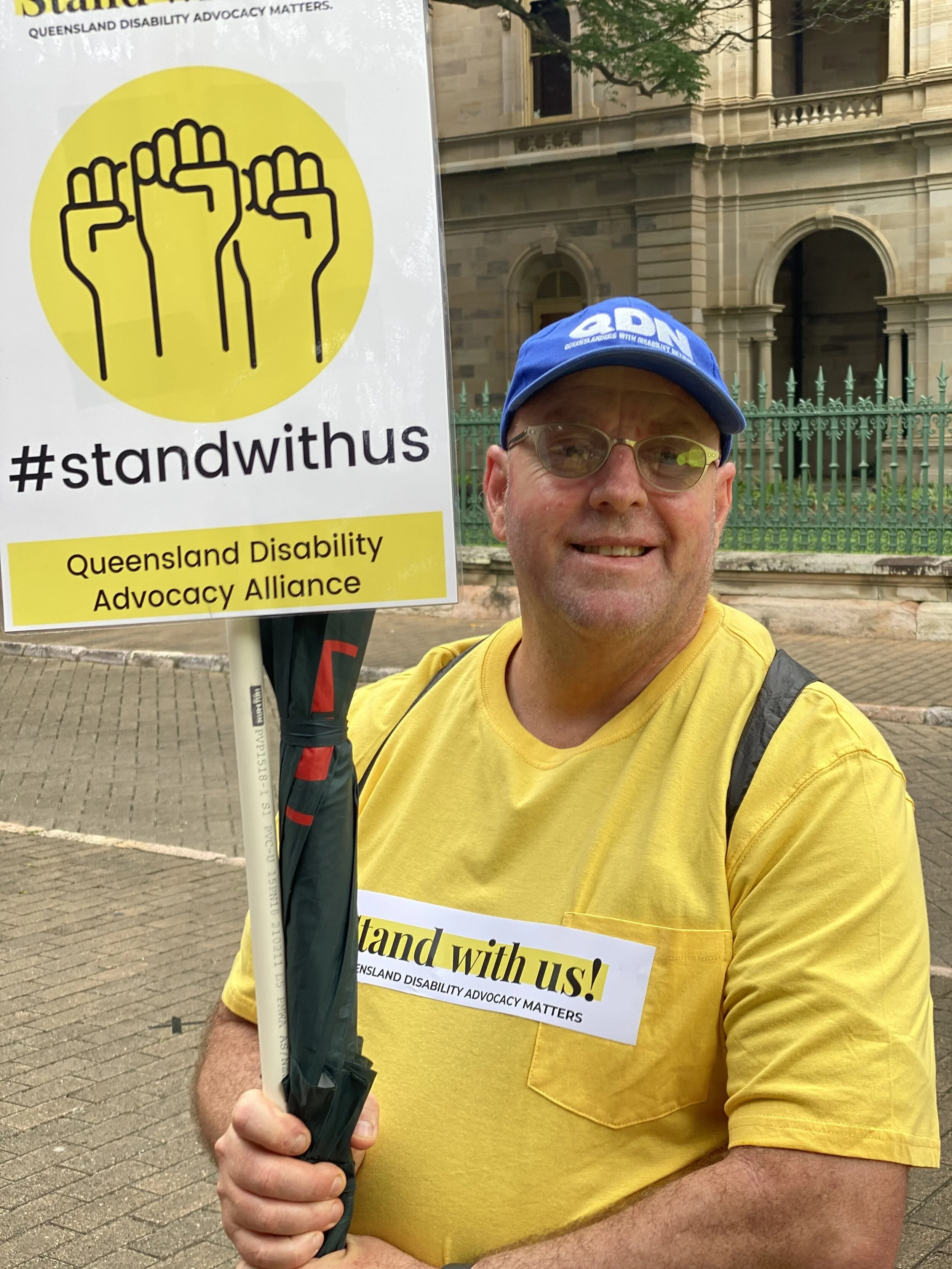 A man wearing a yellow shirt and a blue cap holding a Stand with us sign