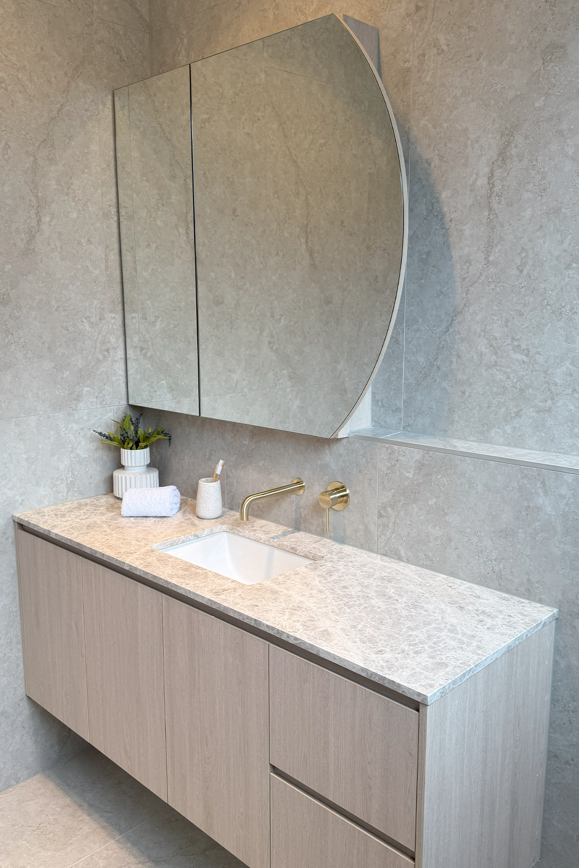 Modern bathroom with a wall-mounted wooden vanity, marble countertop, square white sink, gold faucet, large mirror, small potted plant, folded towel, and toothbrush holder.