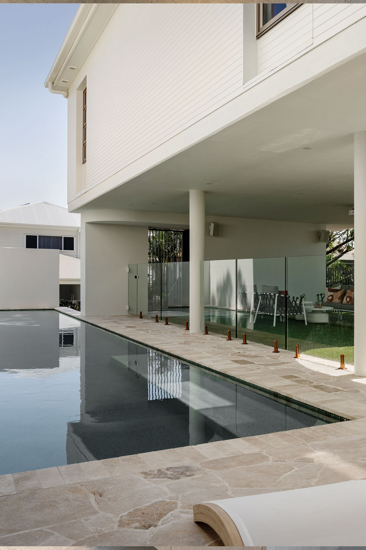 Modern outdoor swimming pool with surrounding patio and glass fencing, adjacent to a contemporary house with overhanging roof.