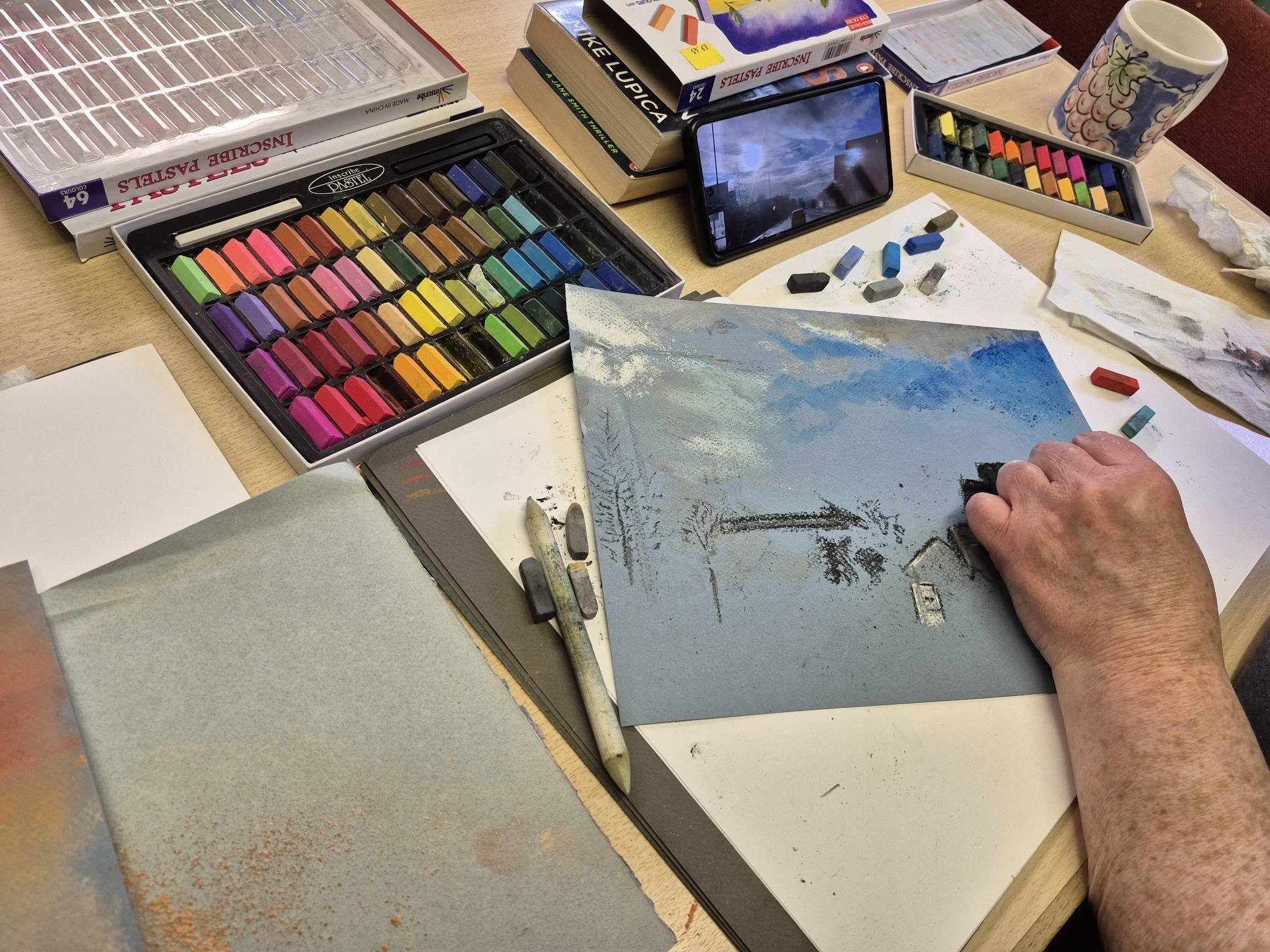 a table with paper and pastels being used. One hand is shown drawing a landscape scene.