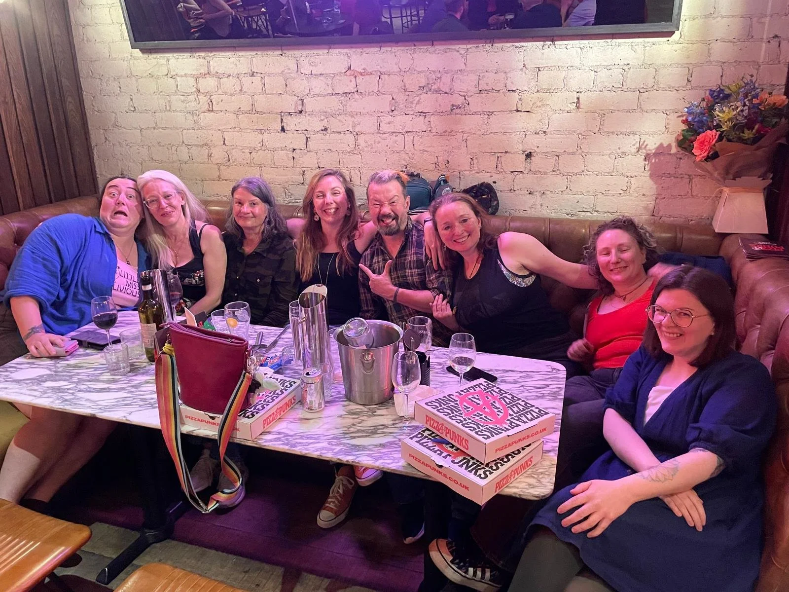 A group of people smiling around a table with various glasses and pizza boxes on it.