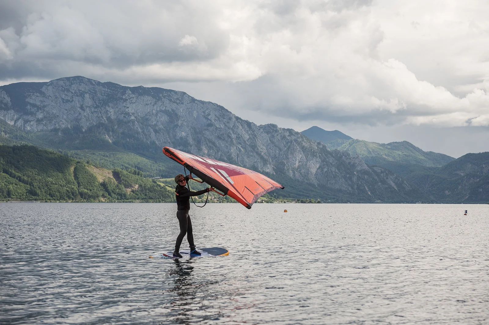 Person steht auf einem Wingfoil Board in einem See, umgeben von Bergen, bei bewölktem Himmel.