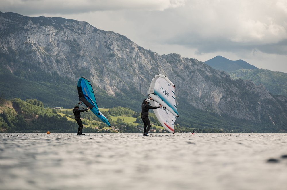 Zwei Wingfoiler auf einem See vor Bergen, die Vorbereitungen zum Wingsurfen treffen.