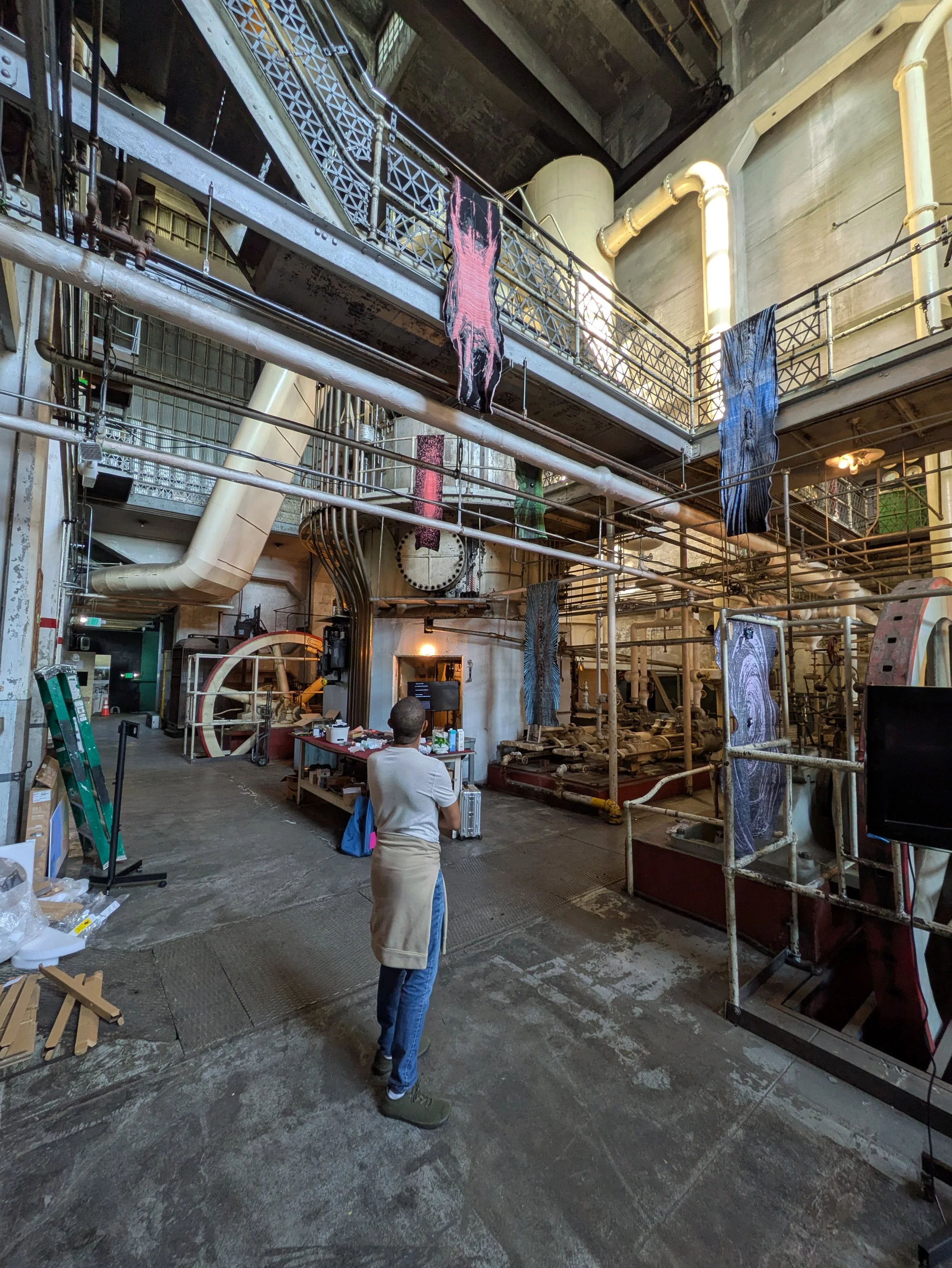 A person stands observing textile art hung from industrial machinery. The setting is spacious, with a mix of metal structures and pipes.