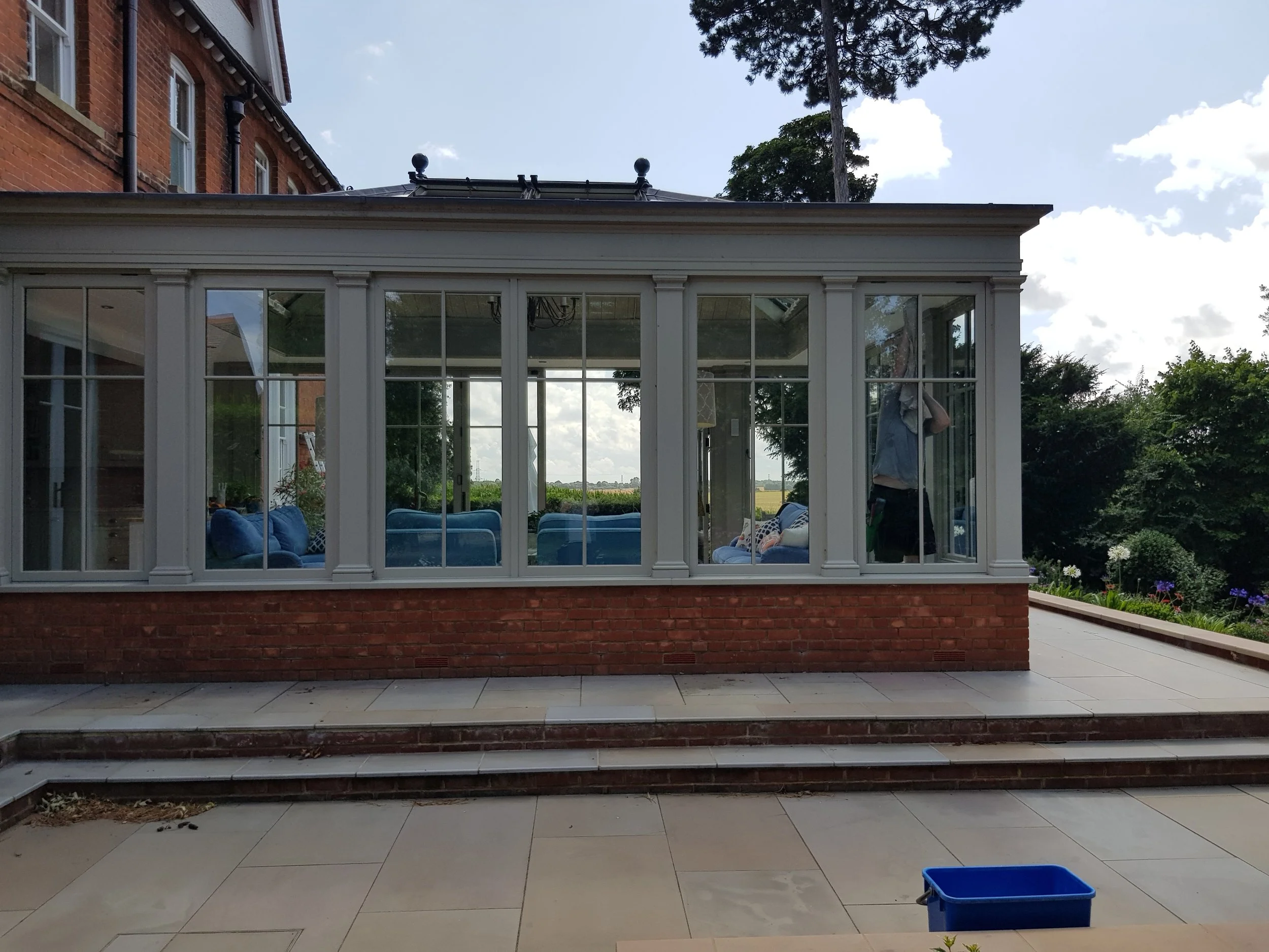 View of a brick house with a large white-framed glass conservatory, with visible interior furniture. A person is partially visible inside, cleaning the conservatory windows. There is greenery and a partly cloudy sky in the background.
