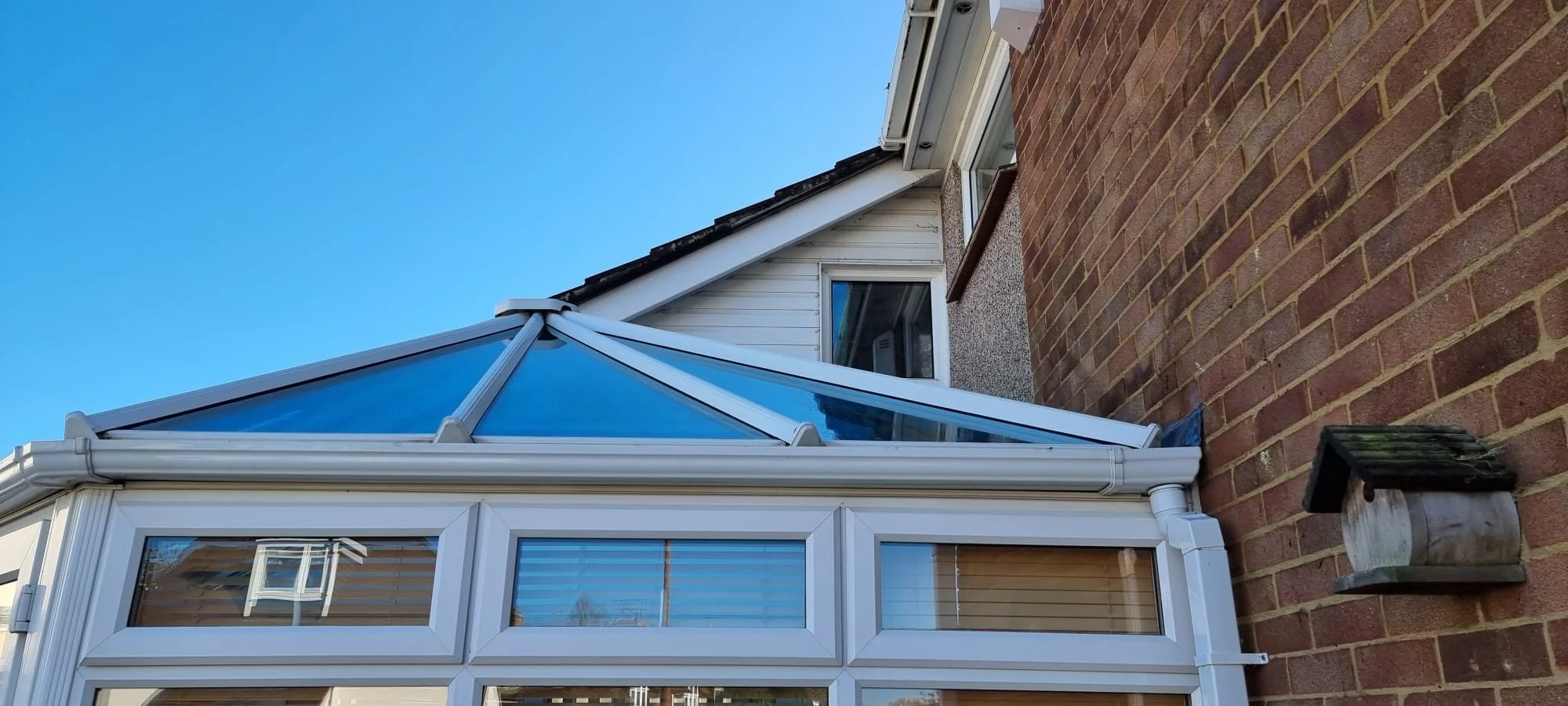 A close-up of a conservatory with a white frame and glass panels attached to the side of a brick house. The conservatory has a sloped glass roof and a small window with blinds. The background includes a clear blue sky.