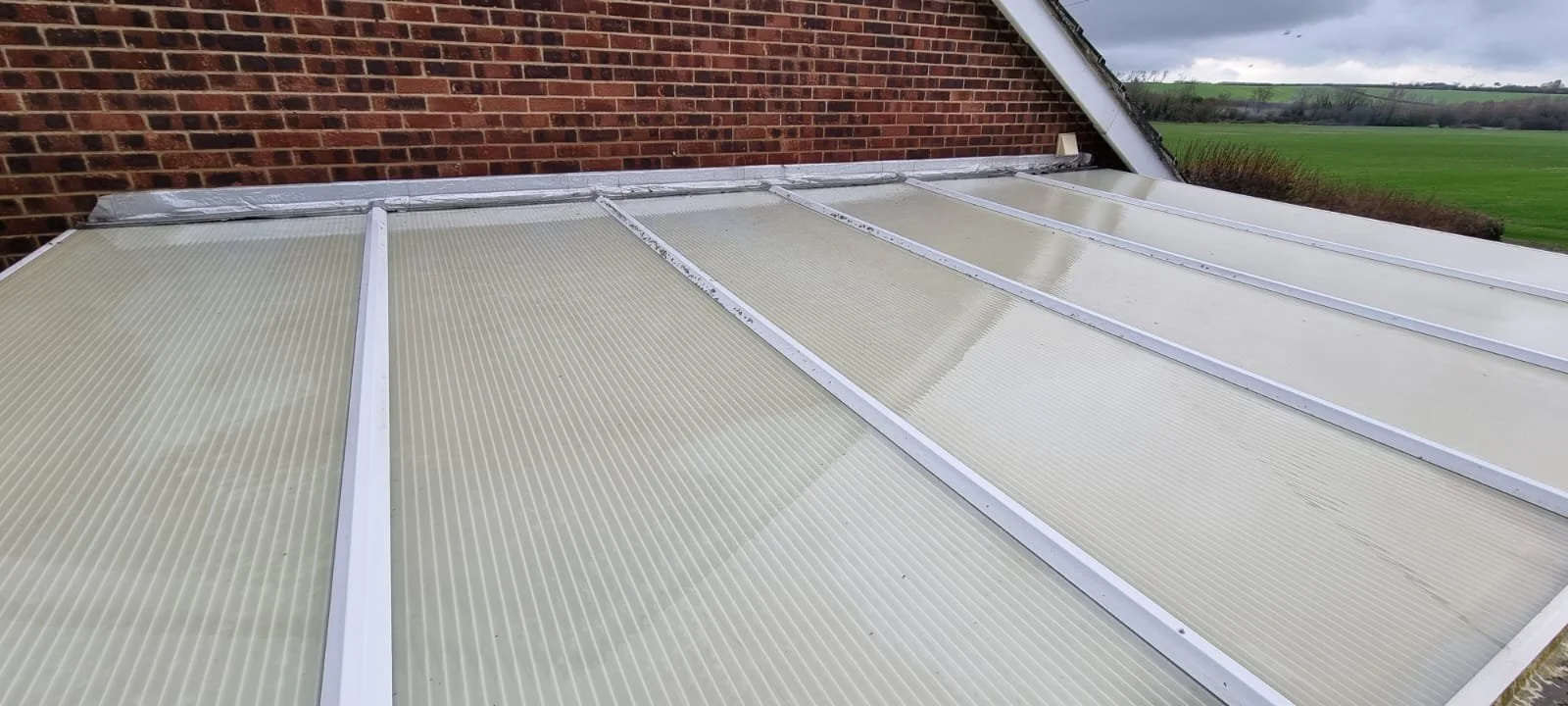 View of a Perspex conservatory roof after cleaning, attached to a brick building with a field and cloudy sky in the background.