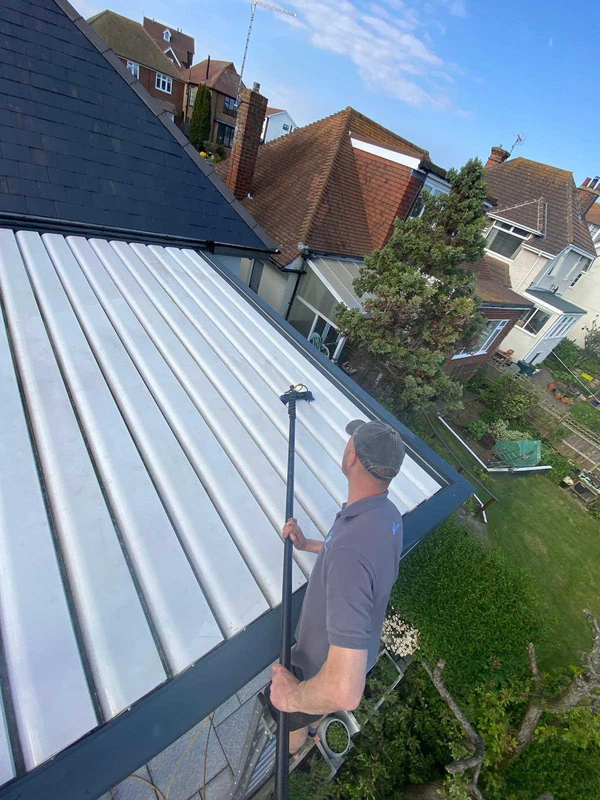 A man standing on a ladder cleaning a sun room roof with a long-handled brush in a residential neighbourhood.