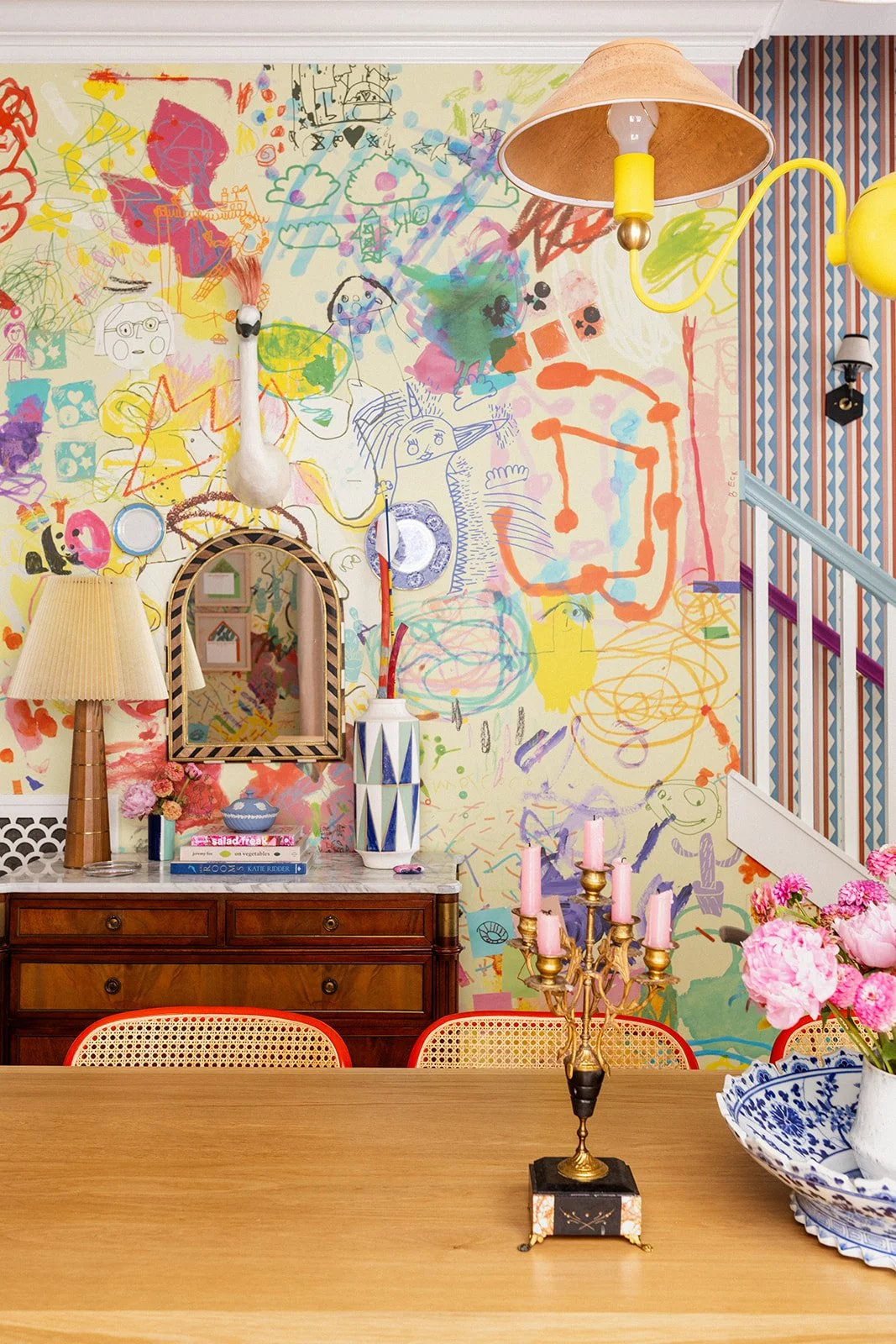 Dining room wall covered in custom wallpaper made from children's artwork, with an eclectic mix of vintage decor, whimsical lighting, and playful sculptural elements.