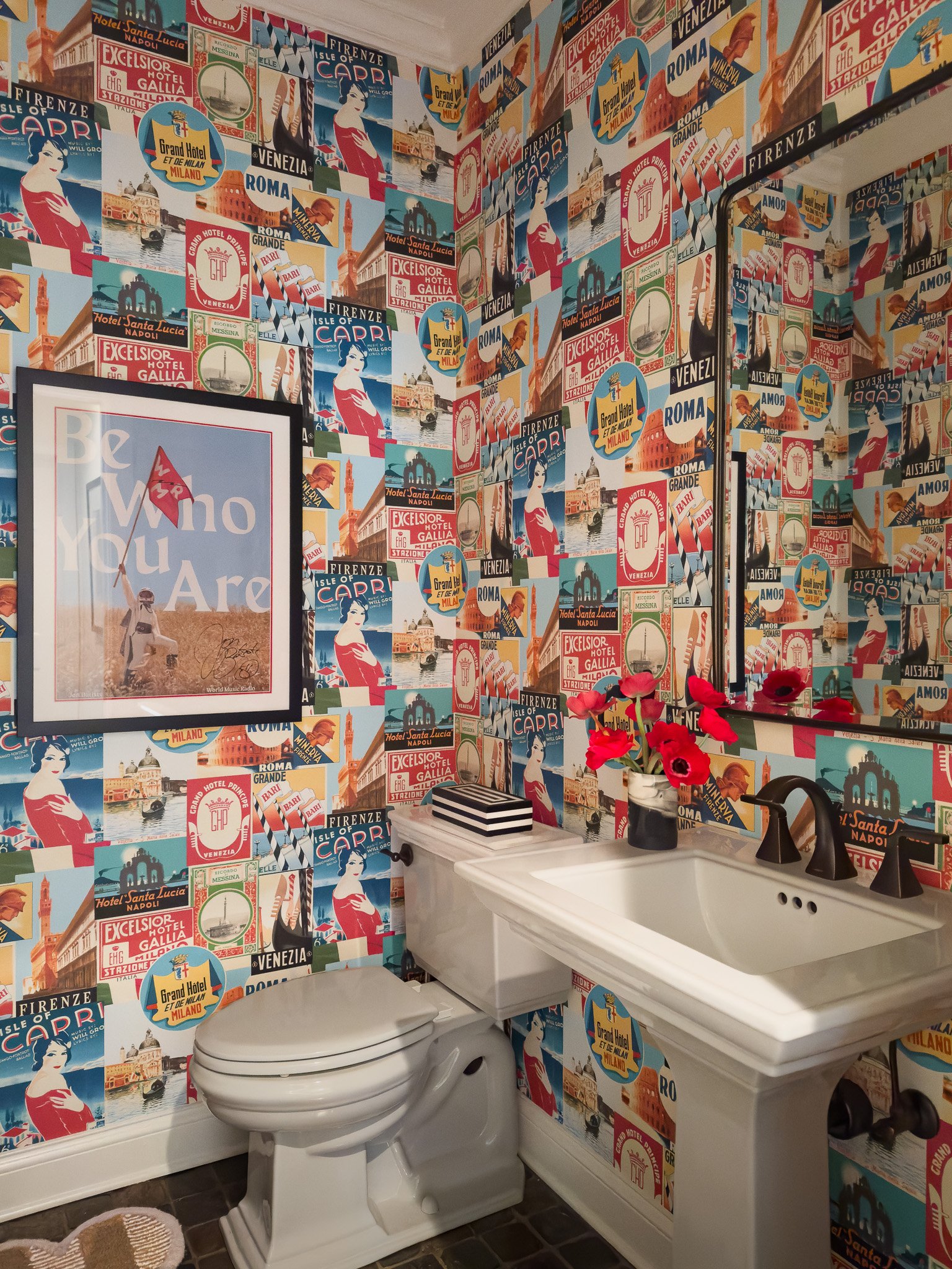 Maximalist powder room wrapped in vintage travel poster wallpaper with pedestal sink, graphic art, and playful red accents — bold design for a tiny space.