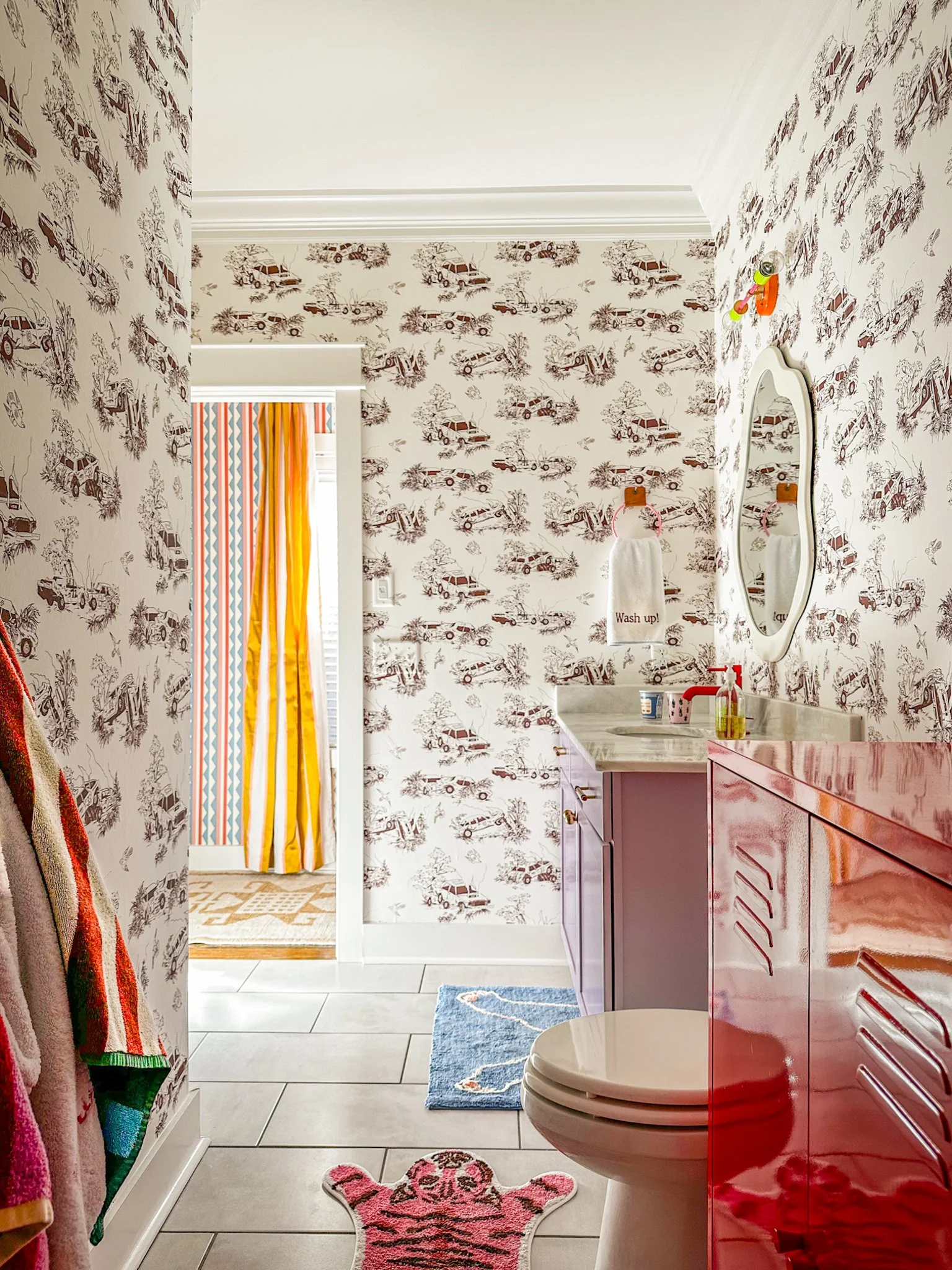 A charming children’s bathroom with playful demolition-derby car-themed toile wallpaper, lavender cabinetry, and colorful bath mats—crafted for high style and high function.