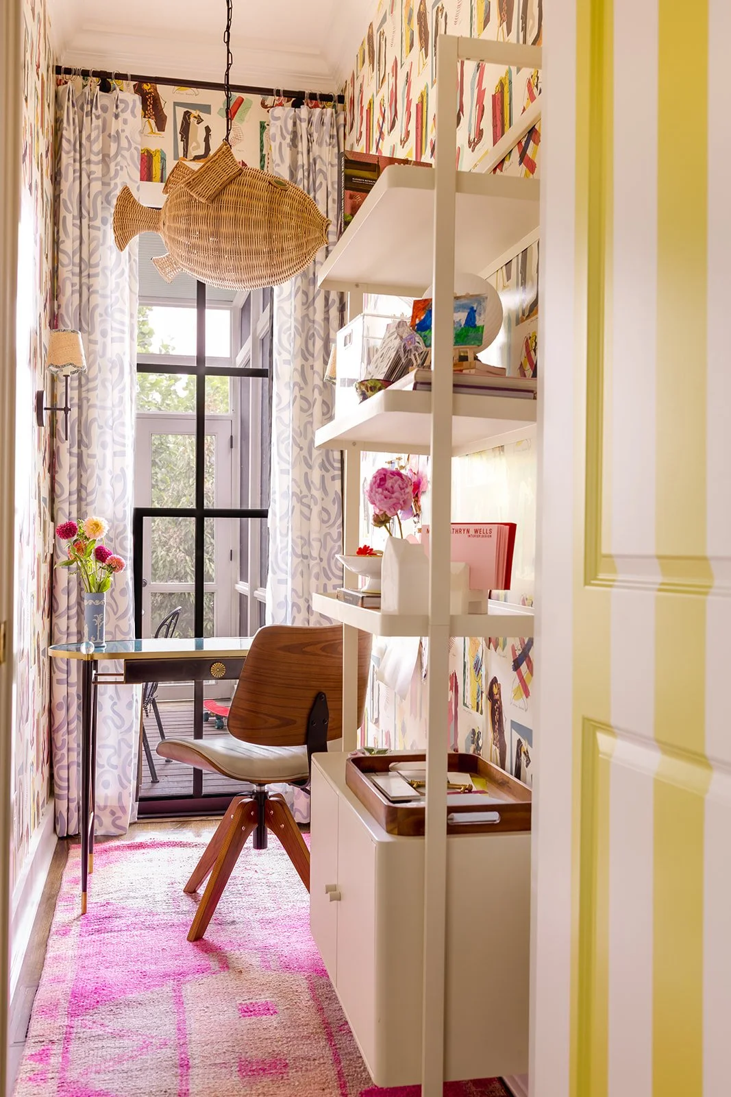 A tiny but bold, wallpapered home office bursting with color and creativity, complete with built-in storage, rattan fish light fixture, and a pink Moroccan-style rug underfoot.