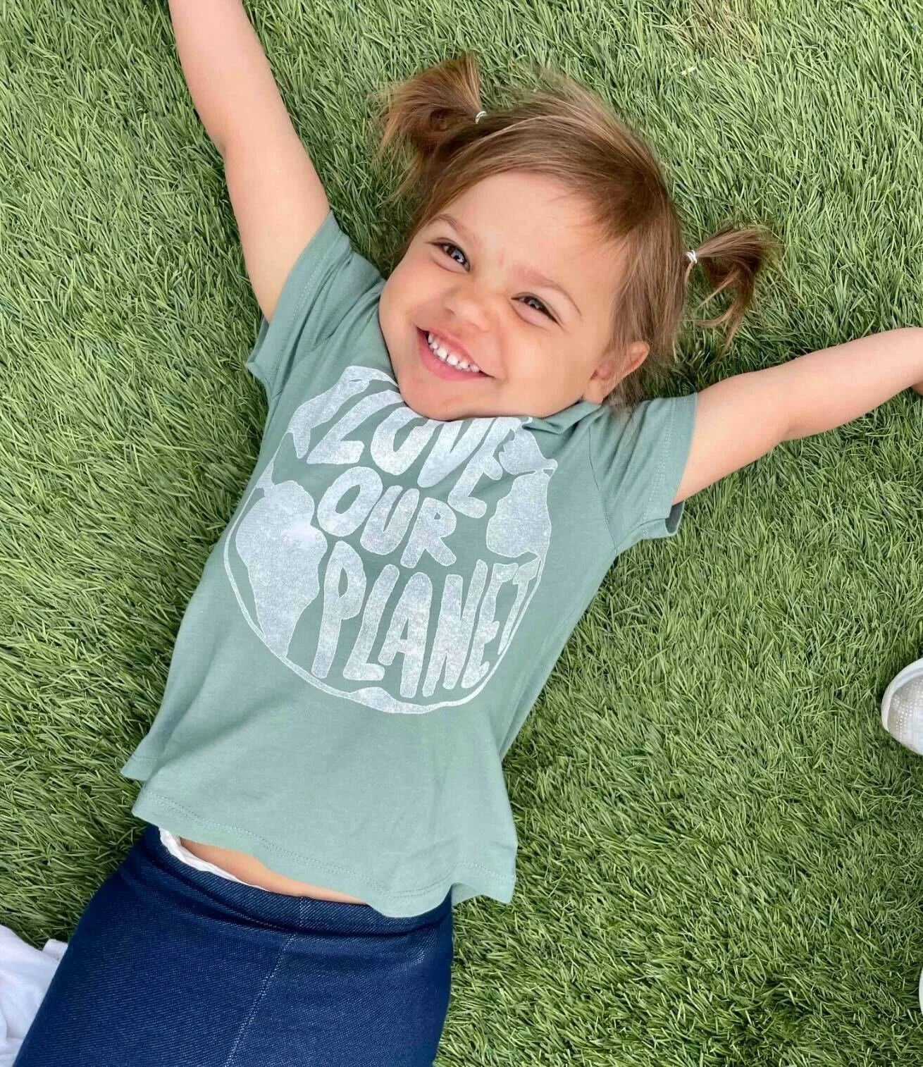 A young girl with light brown hair styled in two pigtails, lying on green grass with arms spread out, smiling at the camera. She is wearing a green T-shirt with the phrase "Love Your Planet" and a graphic of Earth, and dark pants.