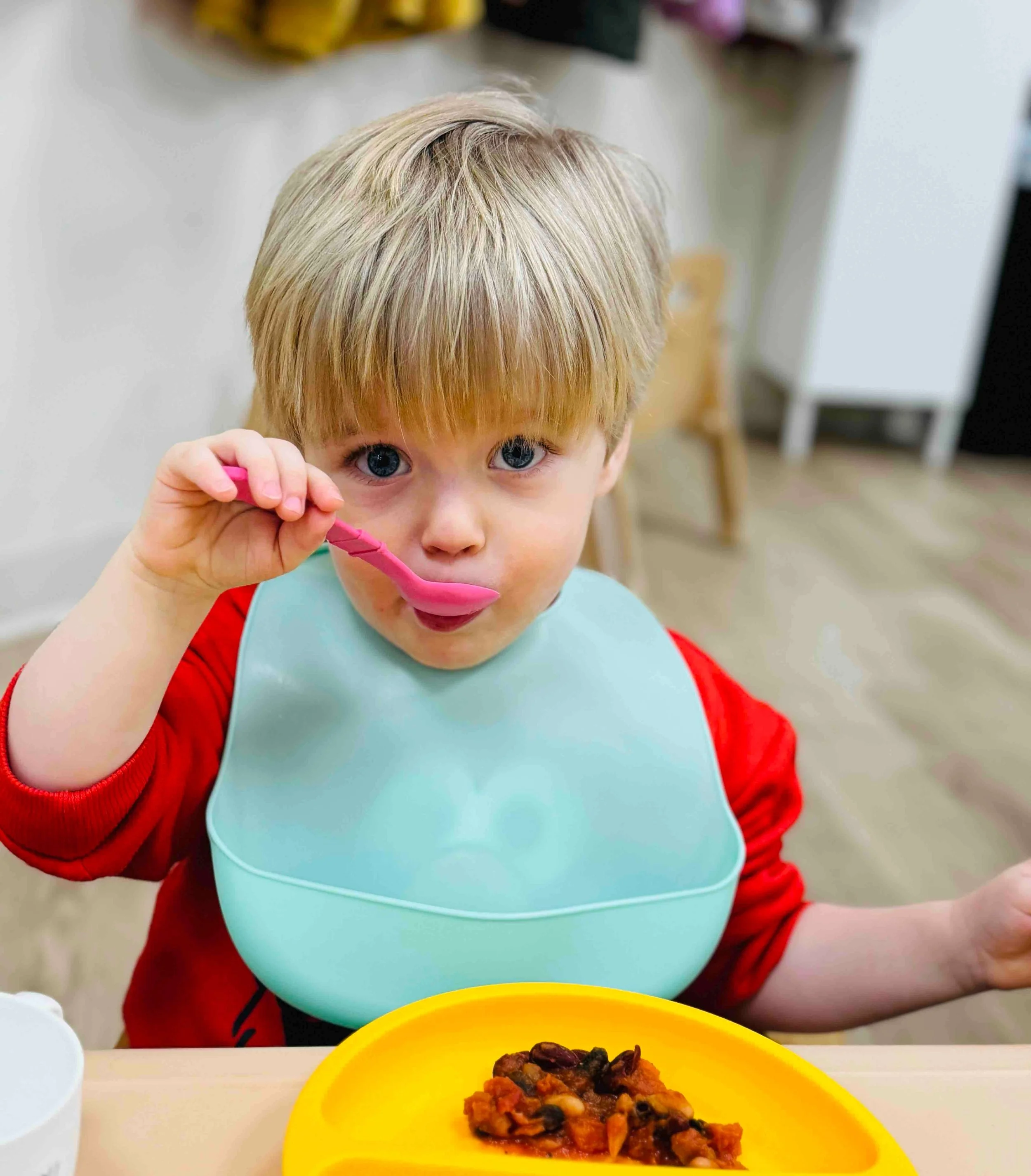 A young boy with blonde hair and blue eyes sitting at a table, holding a pink spoon near his mouth, with a yellow plate of chili at his side.