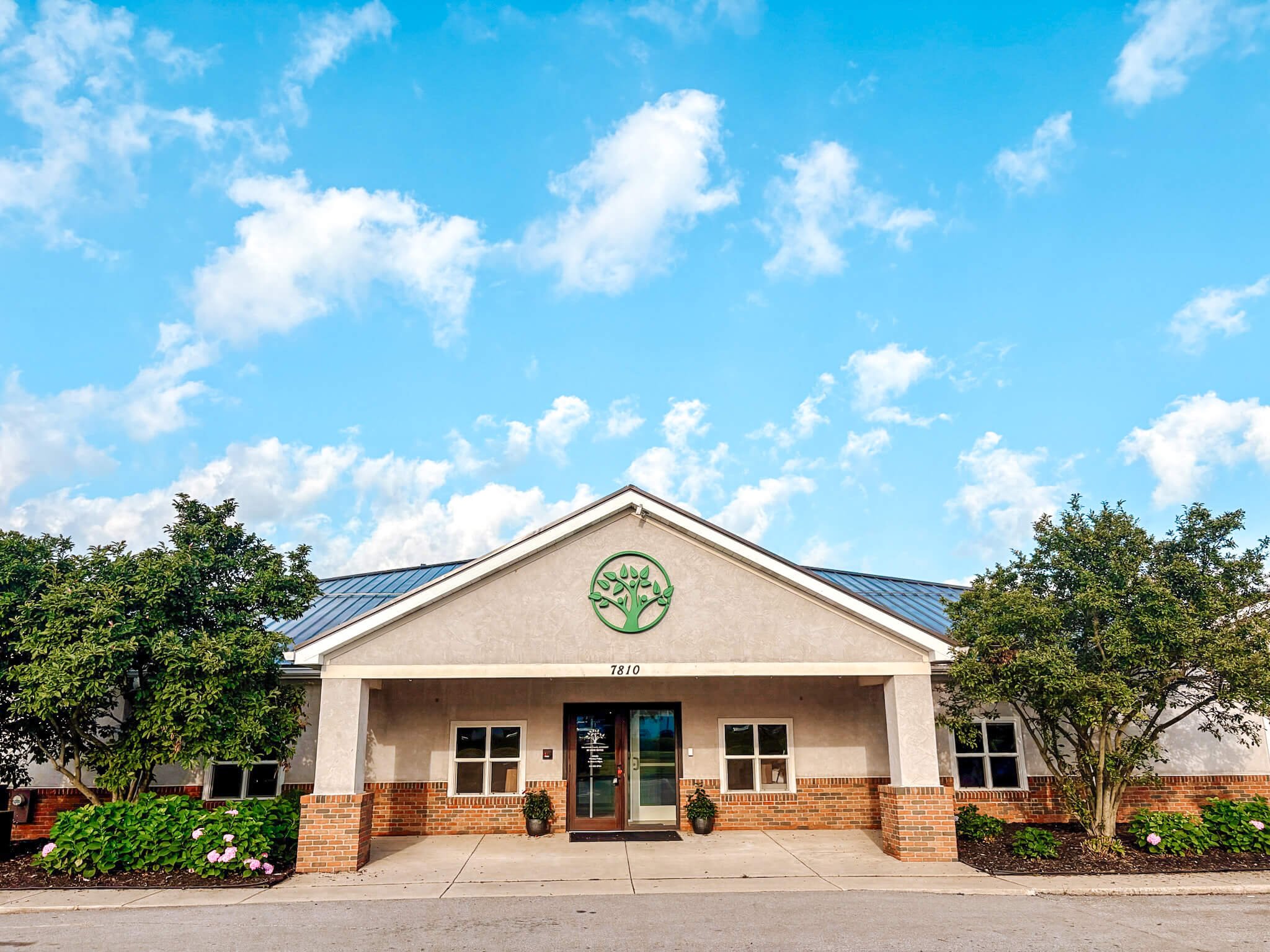 Front view of a building with a tan exterior, brick accents at the base, a porch with columns, and a green circular tree logo above the entrance. Two potted plants flank the door, and trees and shrubs surround the building. The sky is blue with scattered clouds.