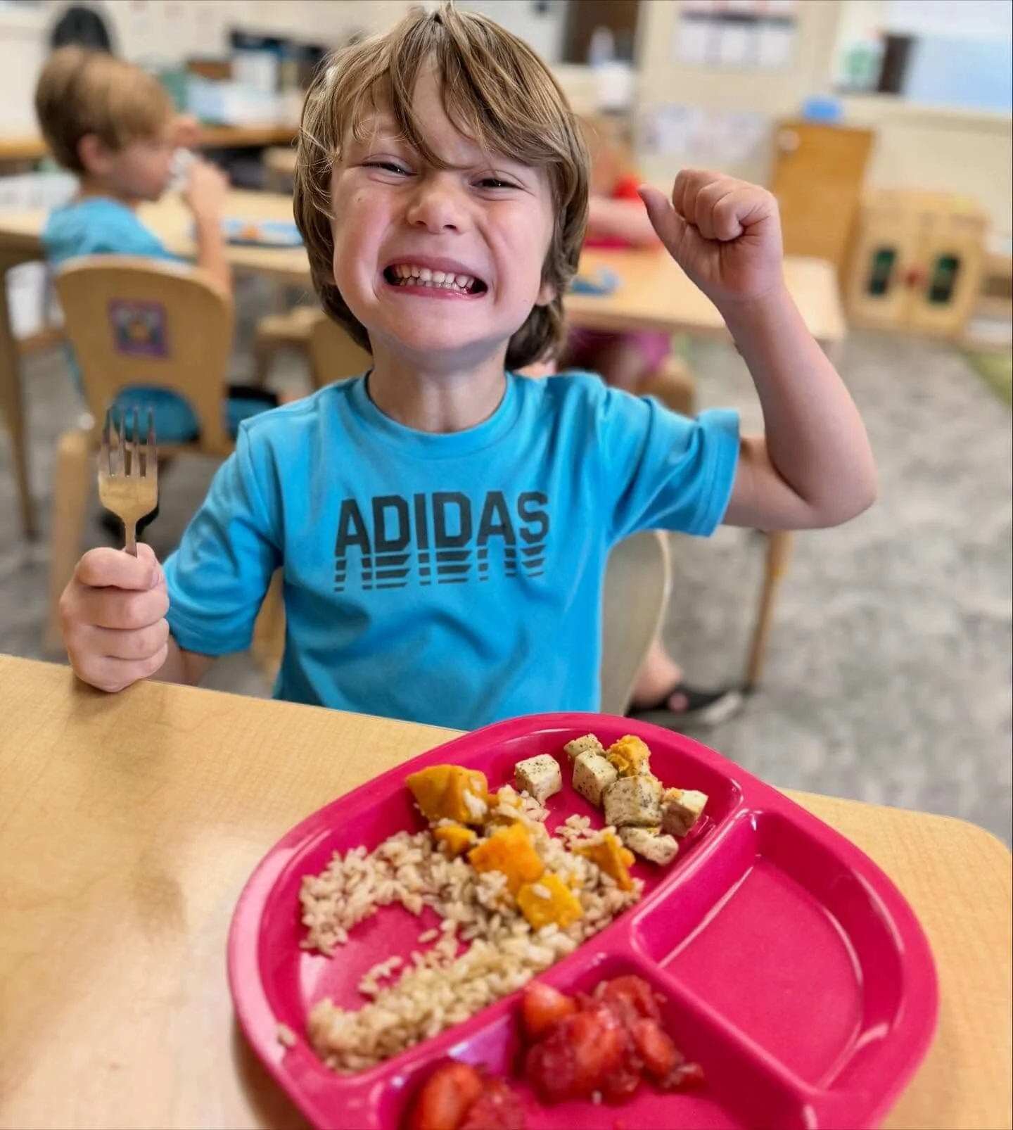 A smiling young boy with light brown hair in a blue Adidas t-shirt, making a fist with his right hand, sitting at a table in a classroom setting. He holds a fork in his left hand, and in front of him is a red divided plate with rice, cubed tofu, and some vegetables. In the background, other children are seated at tables, and classroom shelves are visible.
