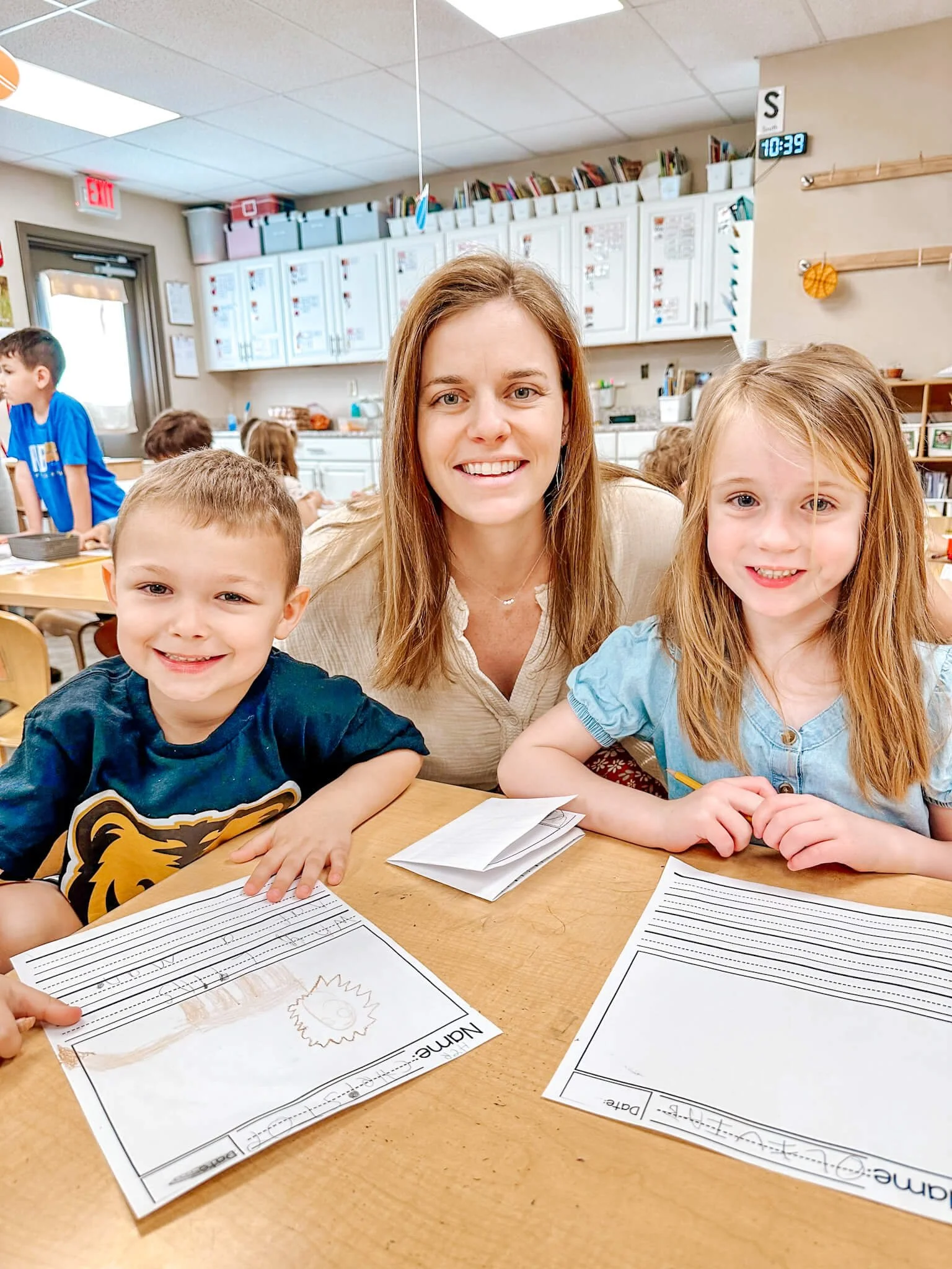 A teacher and two young students smiling at the camera in a classroom, with children and classroom supplies visible in the background.
