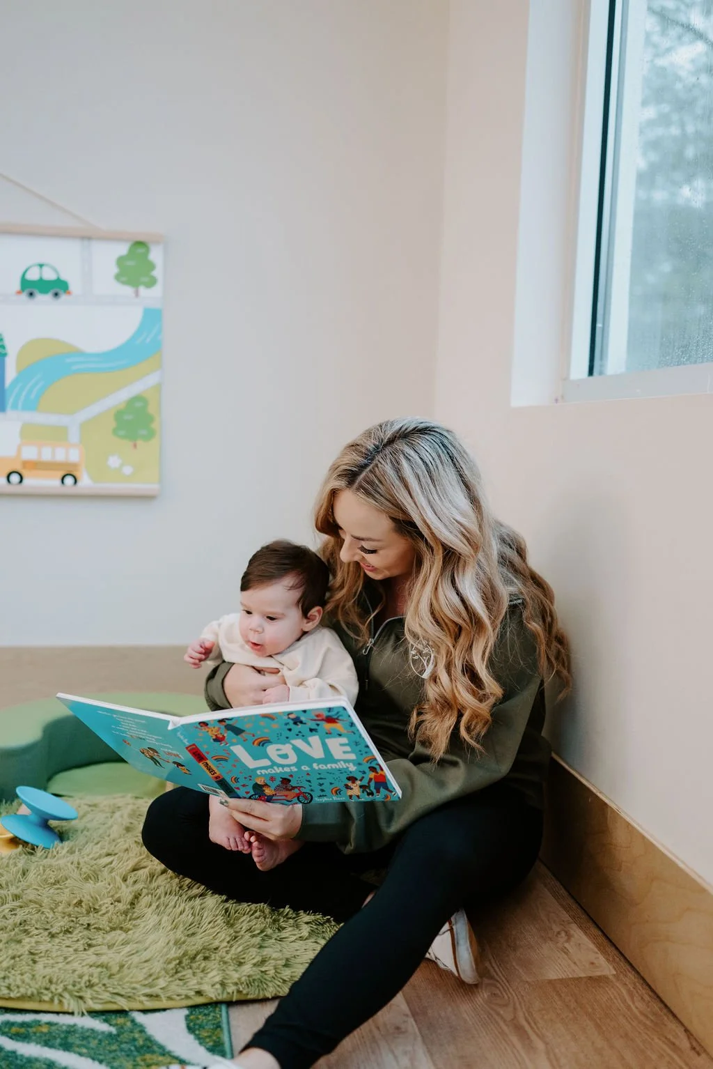 A woman with long, wavy blonde hair sitting on the floor reading a colorful children's book titled 'LOVE makes a family' to a baby sitting on her lap, inside a room with a window and wall art.