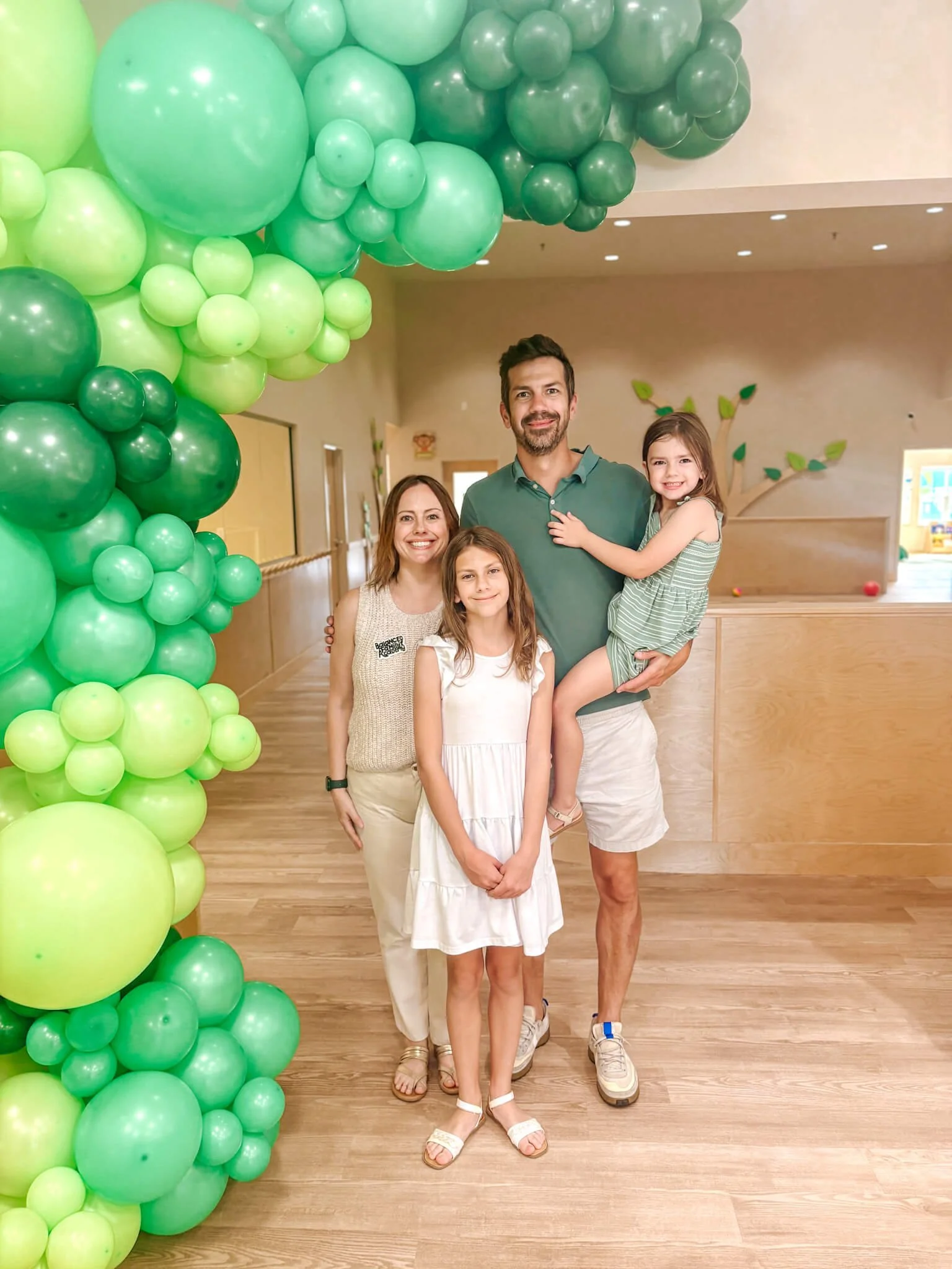 A family of four standing in front of a green balloon decoration in an indoor space. The family includes a man, a woman, and two young girls, all smiling.