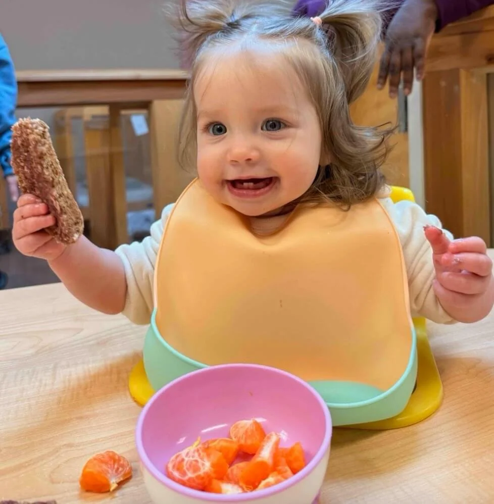 A young girl with brown hair in pigtails wearing a bib, smiling while sitting at a wooden table with a bowl of peeled orange segments and holding a breaded snack.