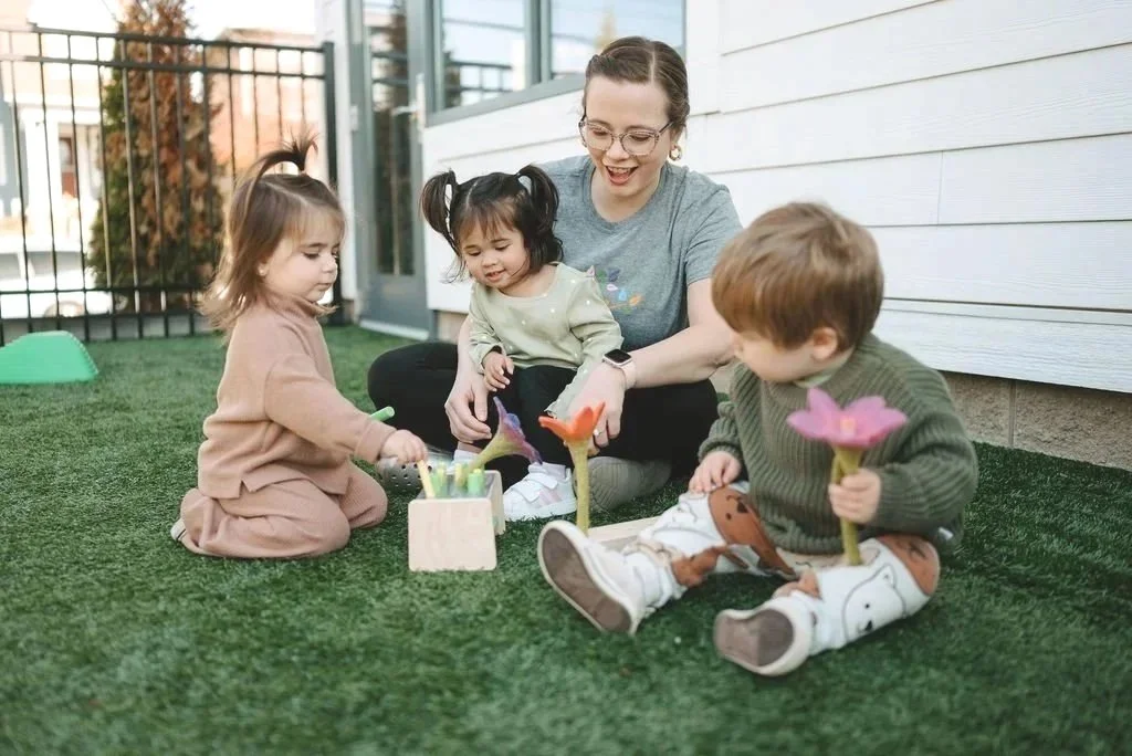 A woman and three children sitting on grass outside, playing with colorful toy flowers.