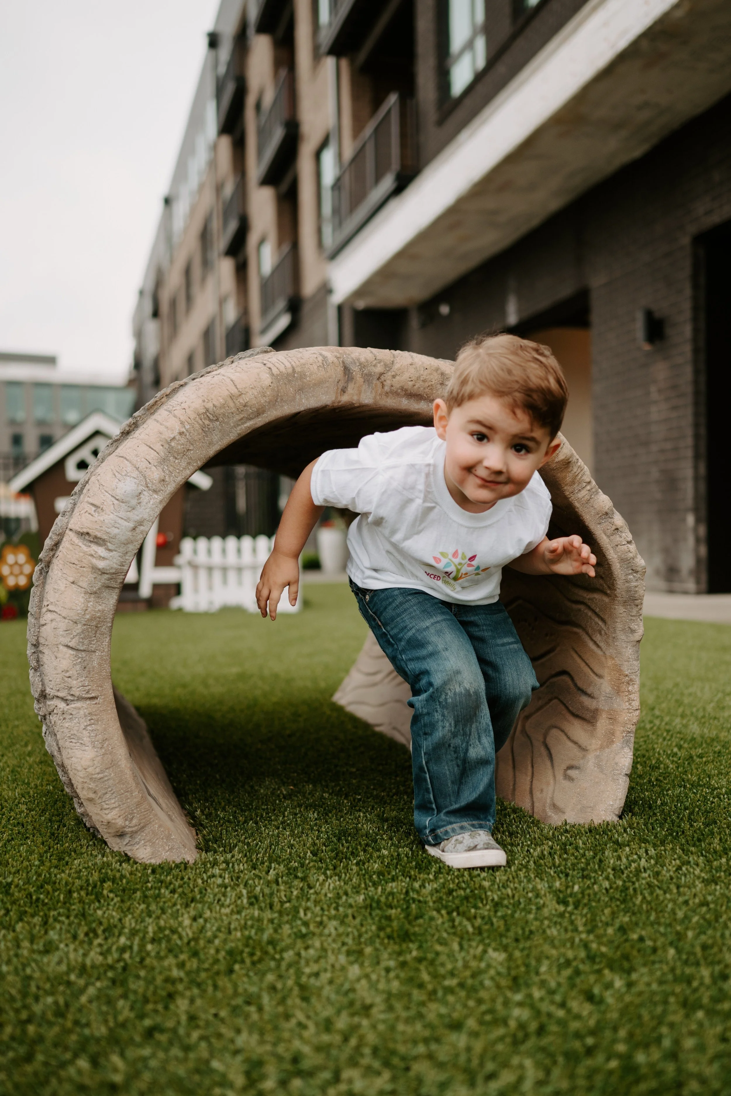 A young boy crawling through a hollowed log in a playground, smiling at the camera with apartment buildings in the background.