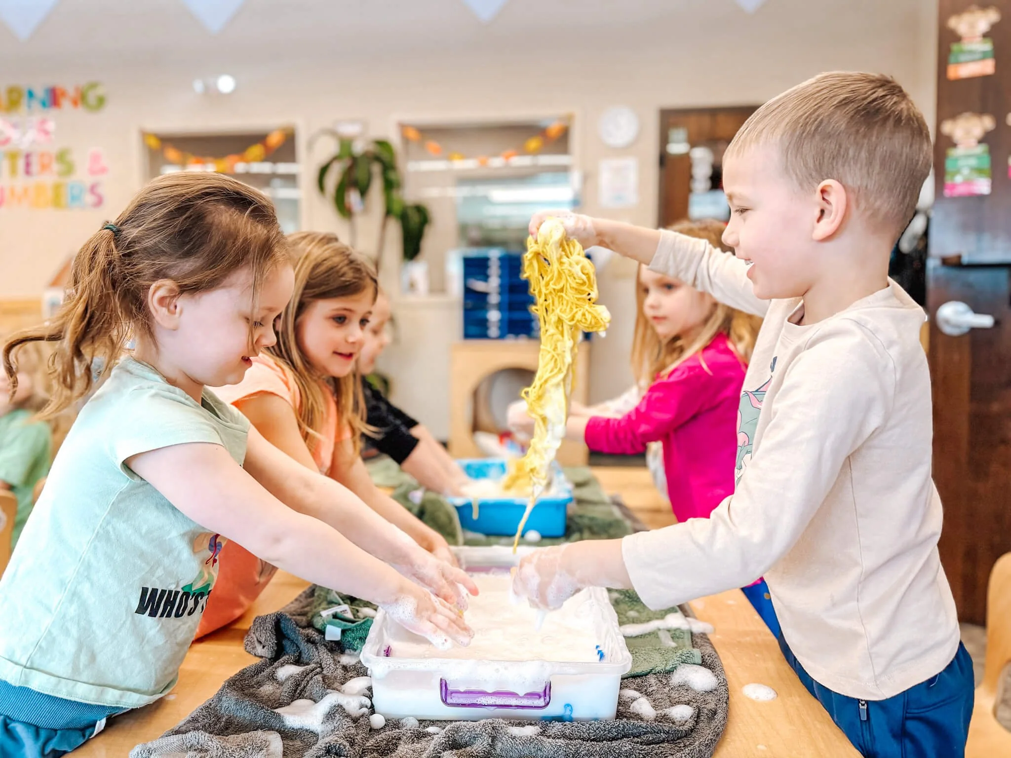 Children in a classroom engaging in a hands-on activity with yellow slime or foam, smiling and interacting with each other at a table.