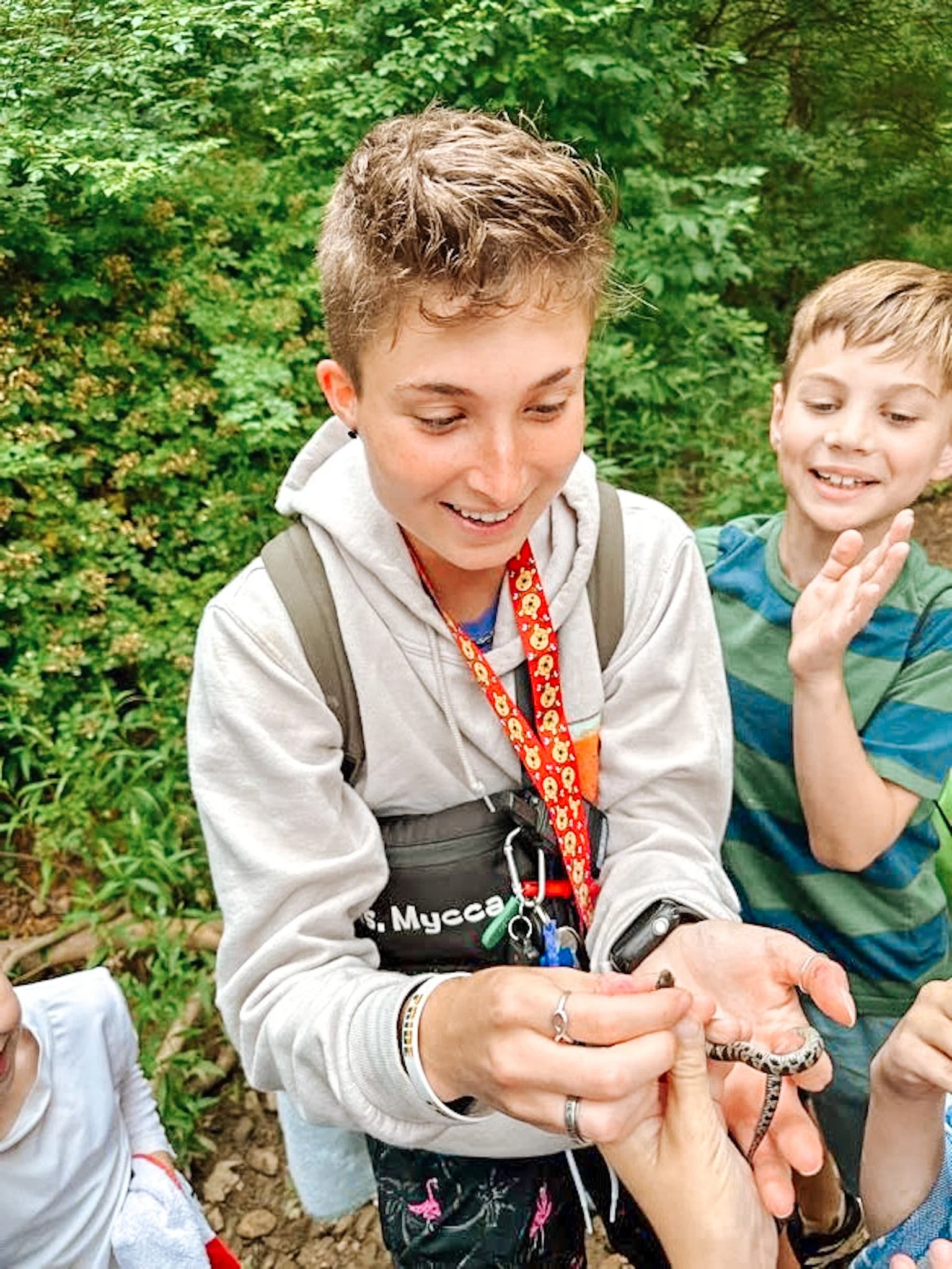 A woman holding a small snake surrounded by children in a green outdoor setting.