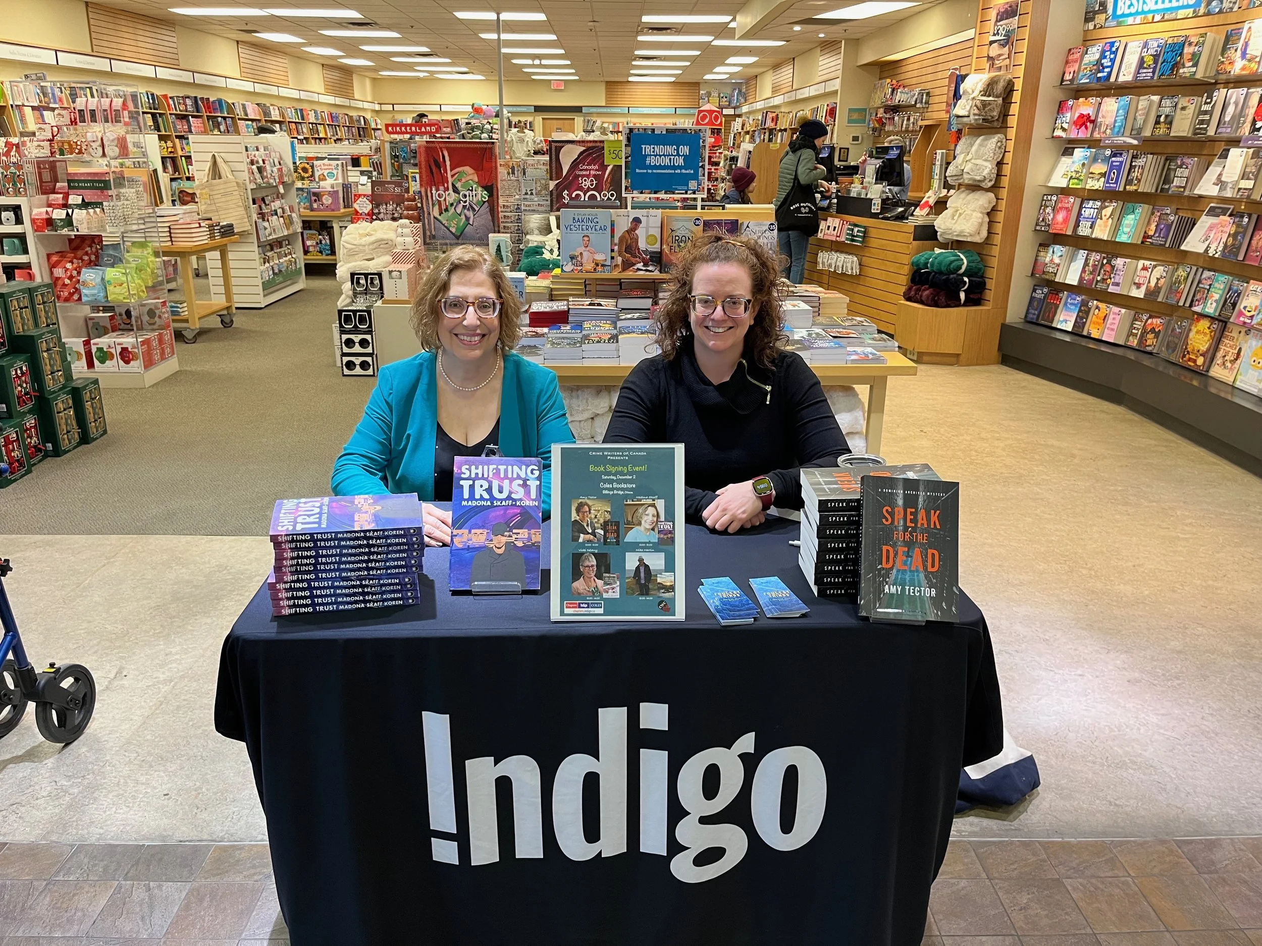 Madona Skaff and Amy Tector behind a table piled with their books, Shifting Trust and Speak for the Dead. Table has a tablecloth covering it, reading Indigo