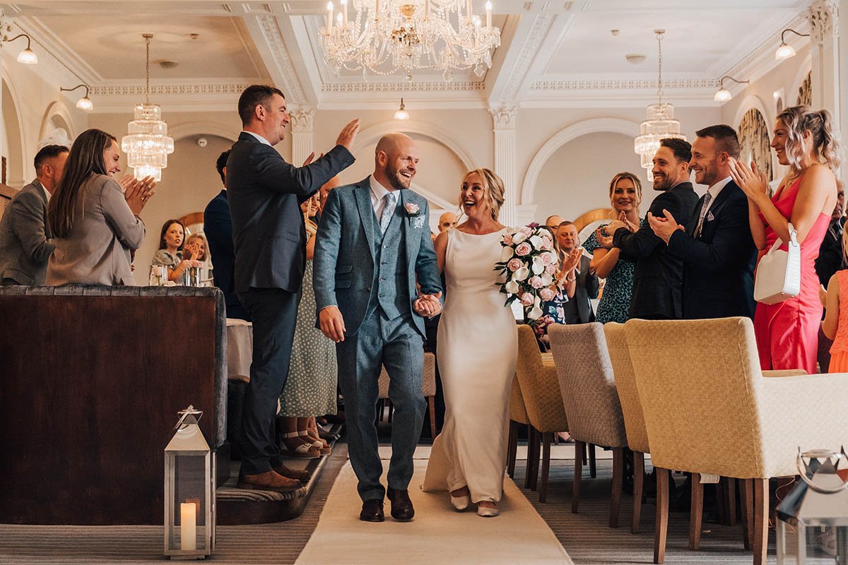A newlywed couple walks down the aisle at The Belsfield Hotel in the Lake District after their wedding ceremony. They hold hands and smile at each other as they make their way down the aisle, with their wedding guests standing on either side smiling