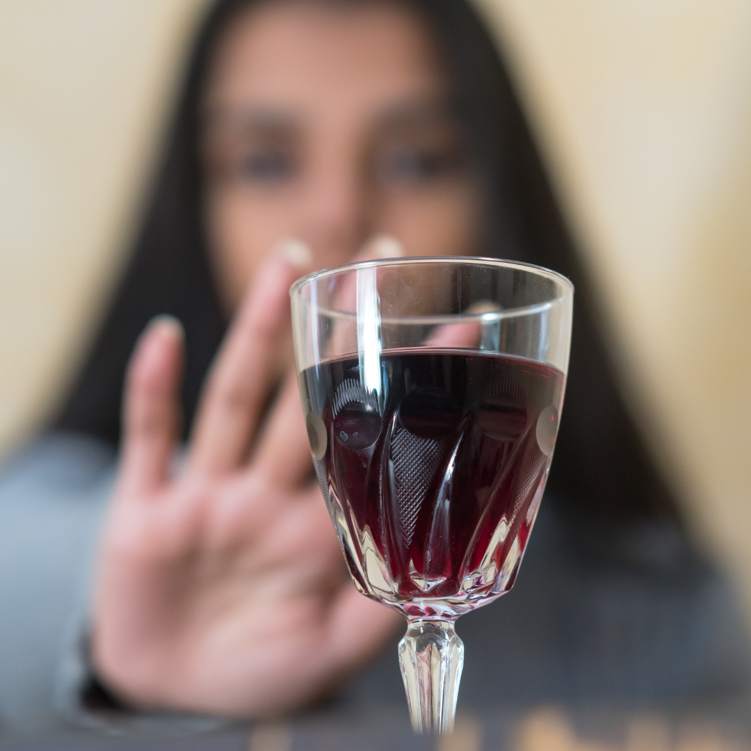 A woman holds her had up to refuse a glass of red wine. She is blurred in the background while the drink is in the foreground of the image.