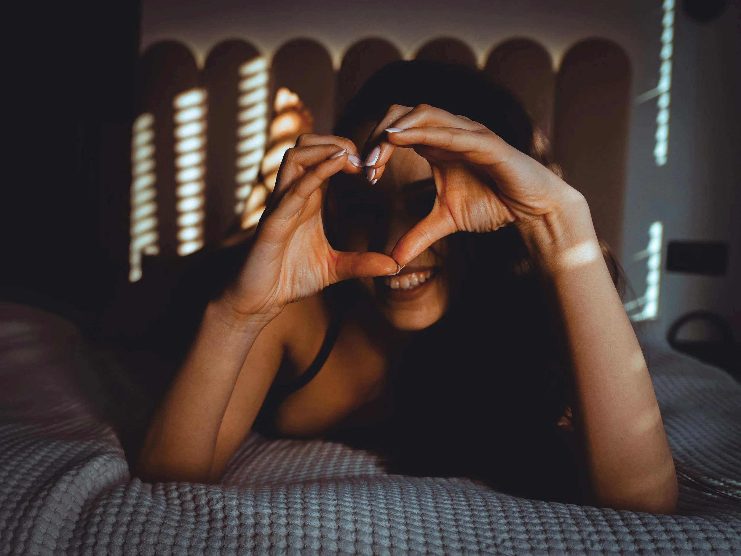 A woman lying on a bed with striped bedding, forming a heart shape with her hands in front of her face, smiling with sunlight streaming through blinds creating shadow patterns.