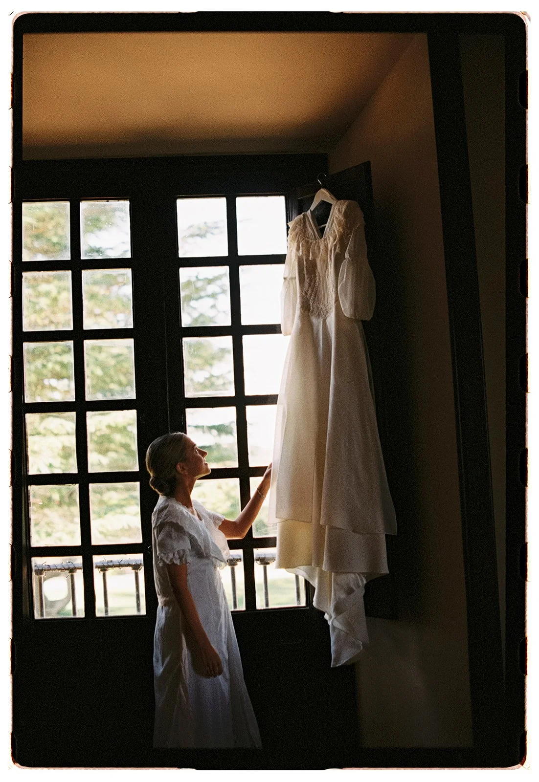 A young girl in a white dress touches a wedding dress hanging on a hook near a window with multiple panes.