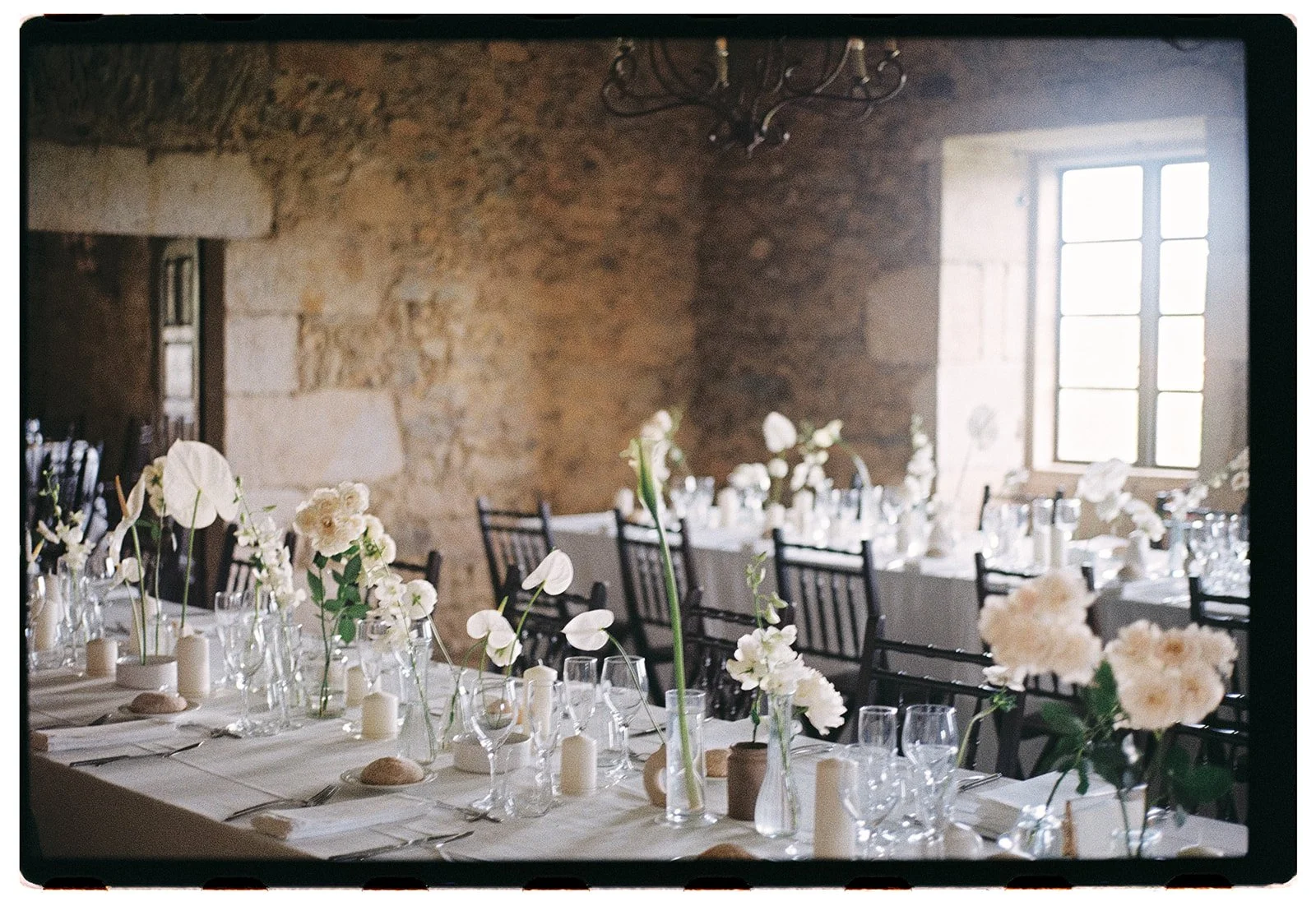 Wedding reception table set with white flowers, candles, glassware, and black chairs in a rustic stone-walled room with a large window.