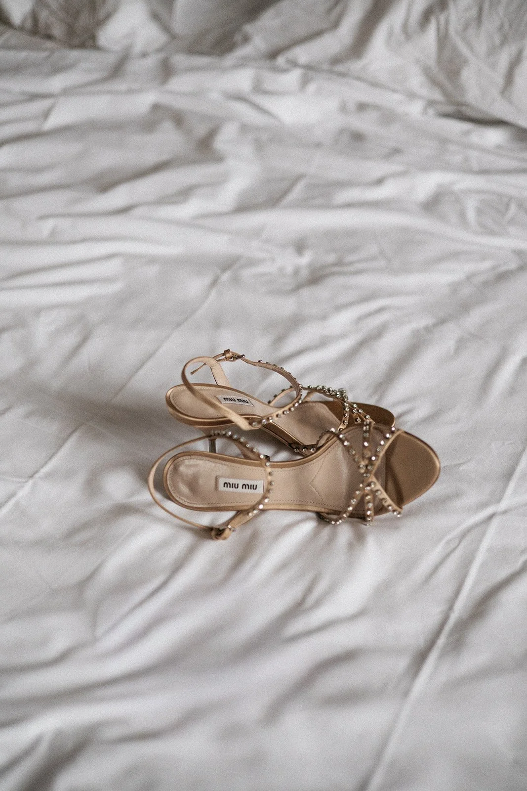 A pair of beige high-heeled sandals with rhinestone straps resting on a white crumpled bedsheet.