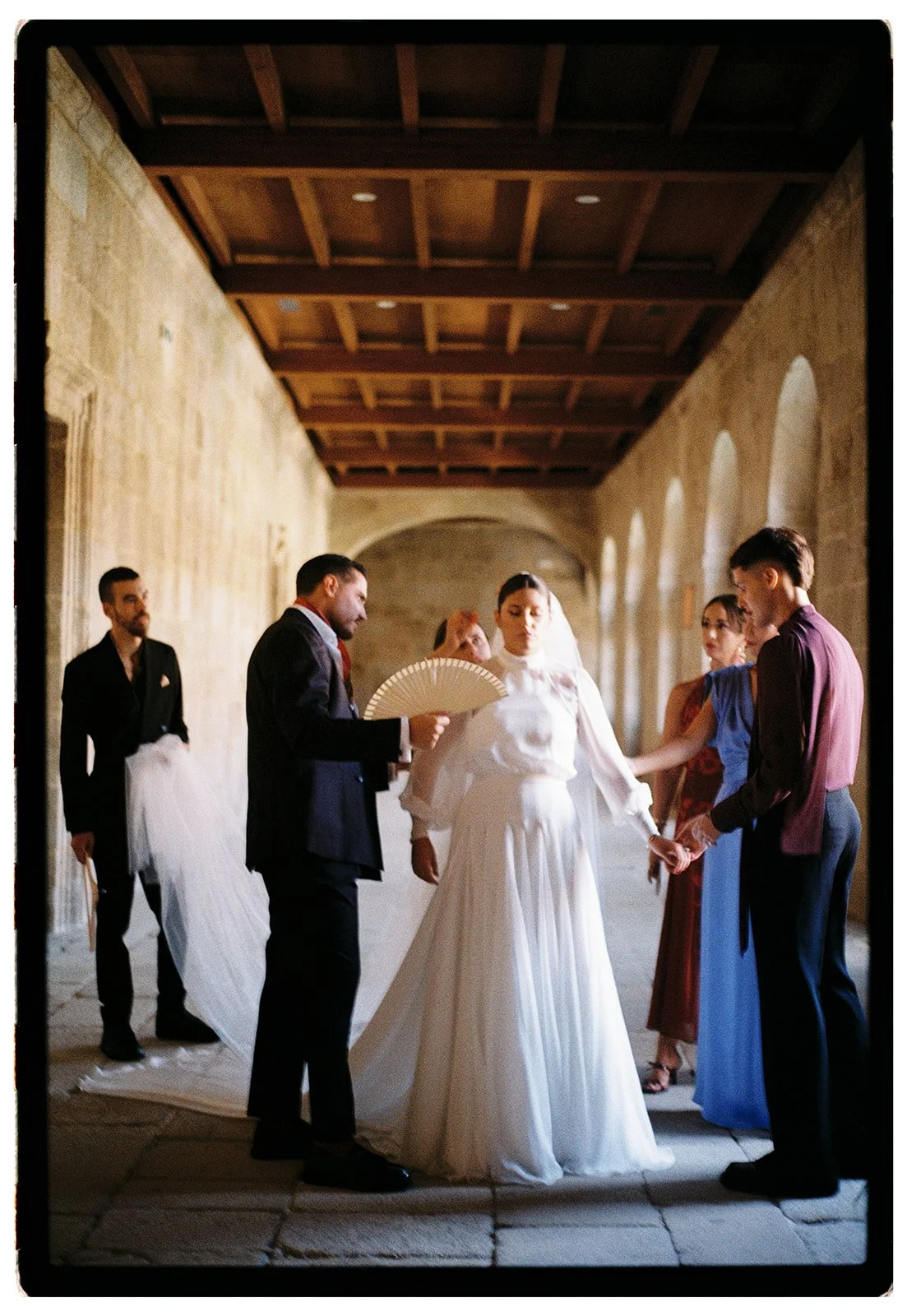 A wedding ceremony with the bride and groom holding hands, surrounded by friends or family in a corridor with stone walls and wooden ceiling.