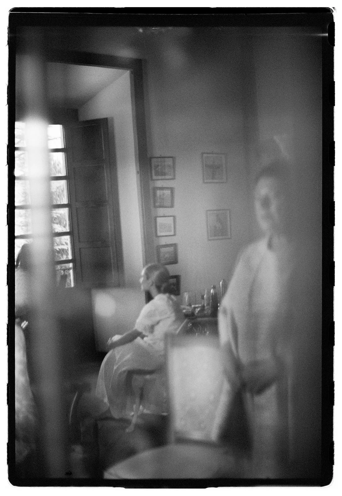 A black-and-white photo showing a woman sitting on a bed in a room with a window, pictures on the wall, and a dresser with bottles and glasses, viewed through what appears to be a mirror or glass surface.