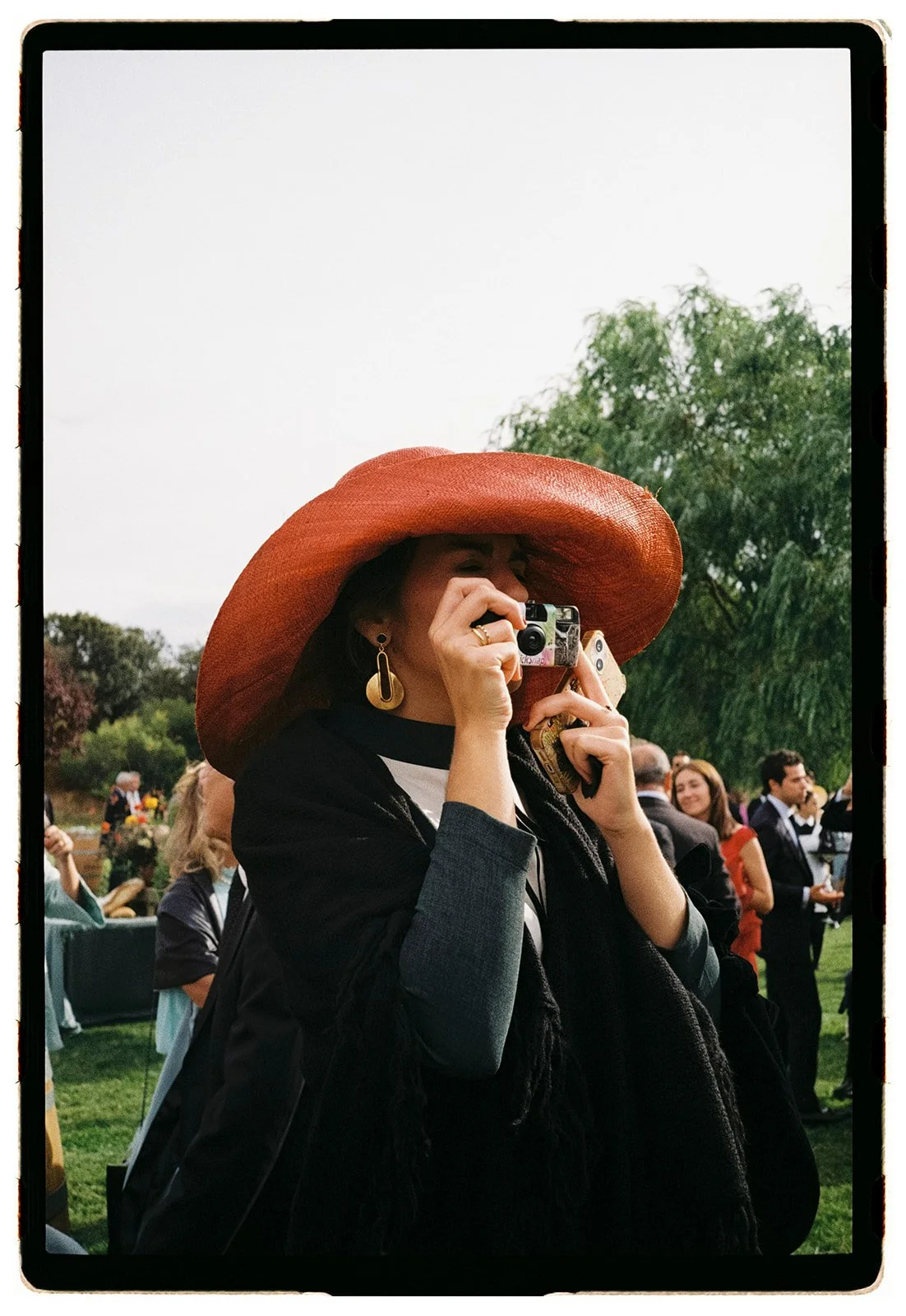A woman wearing a large orange sunhat and black jacket, taking a photo with her camera at an outdoor event.
