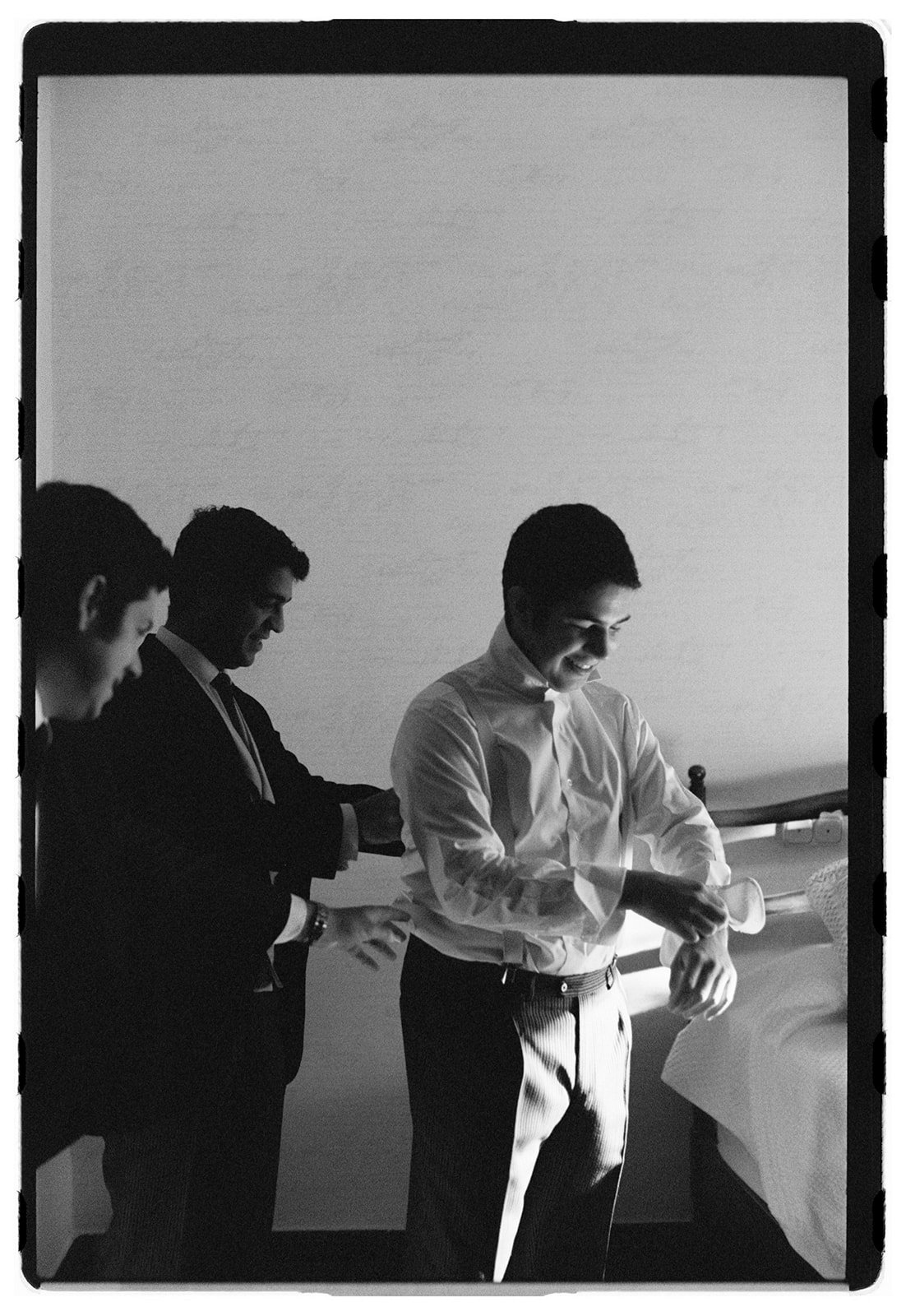 Three men in formal attire standing in a line, smiling, in a room with a bed in the background, with light coming from the right.