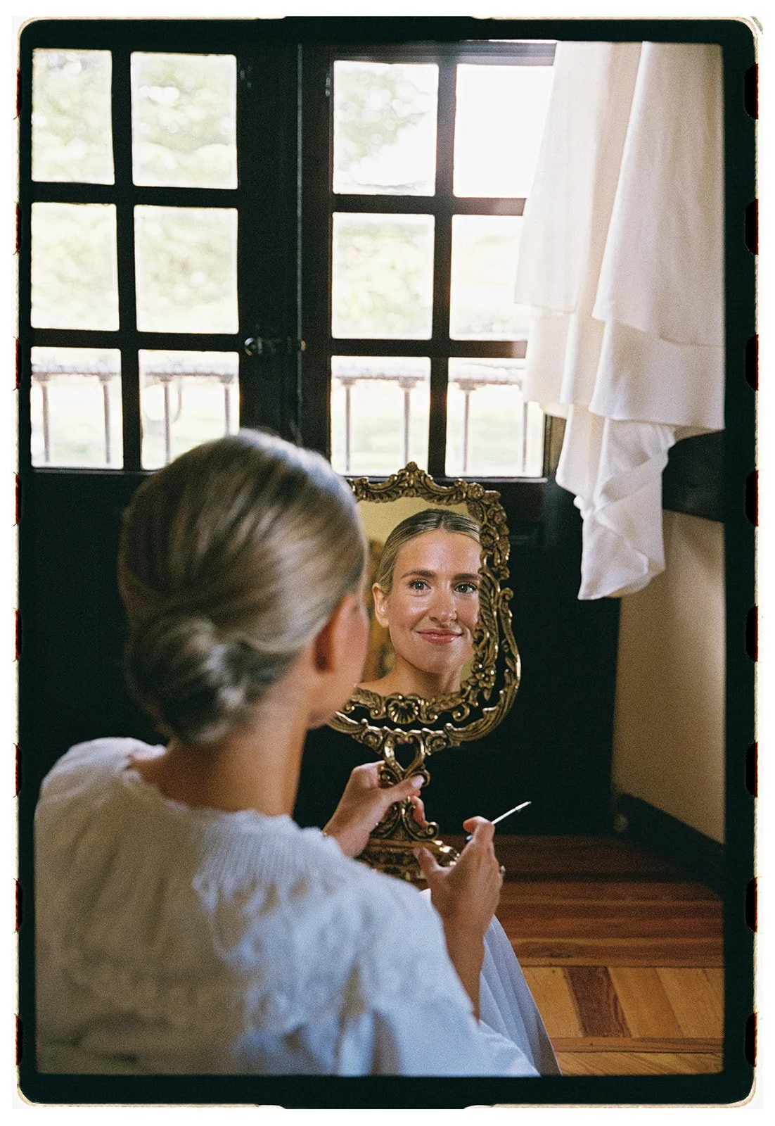 A woman with gray hair in a bun looking at her reflection in a decorative gold-framed mirror, with a window behind her letting in natural light.