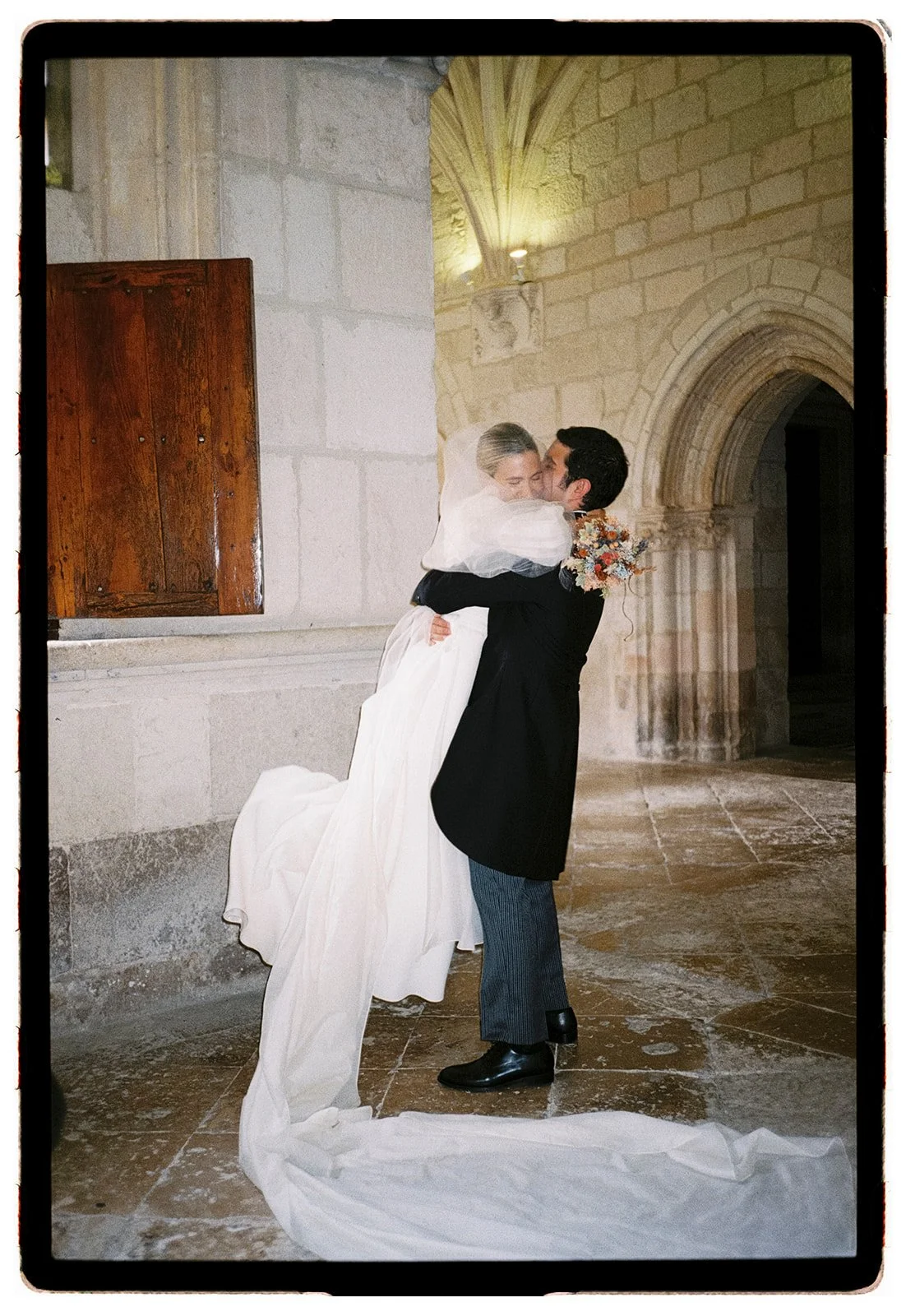 A bride and groom share a kiss inside a historic stone church, with the groom lifting the bride who is wearing a white wedding dress and veil, and the bride holding a bouquet of flowers.