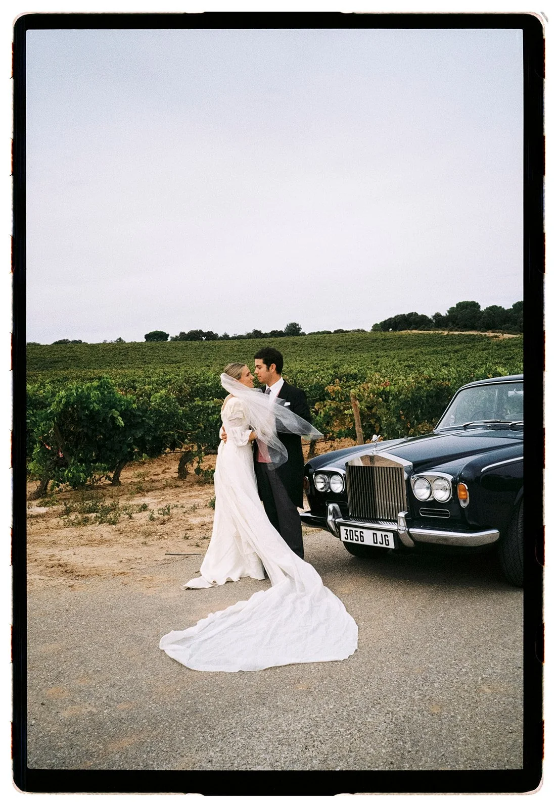 A bride and groom sharing a moment outside next to a classic black car, with a vineyard in the background.