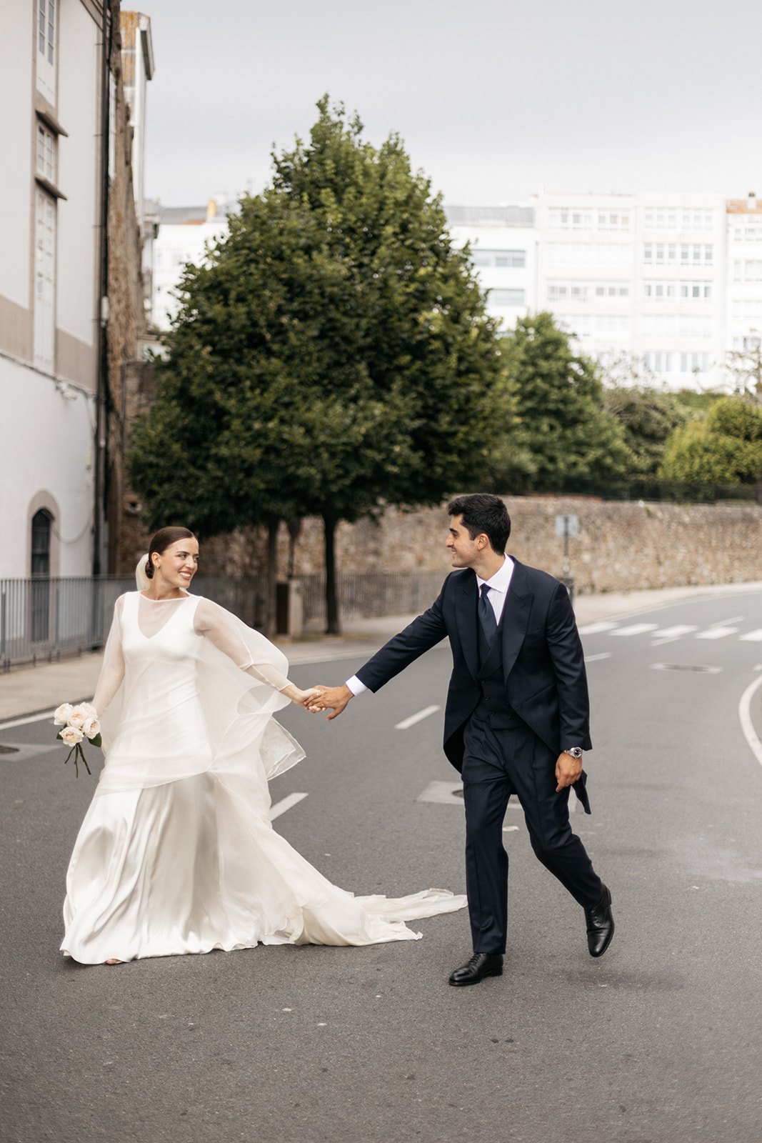 A bride and groom holding hands while walking on a city street, with trees and buildings in the background.