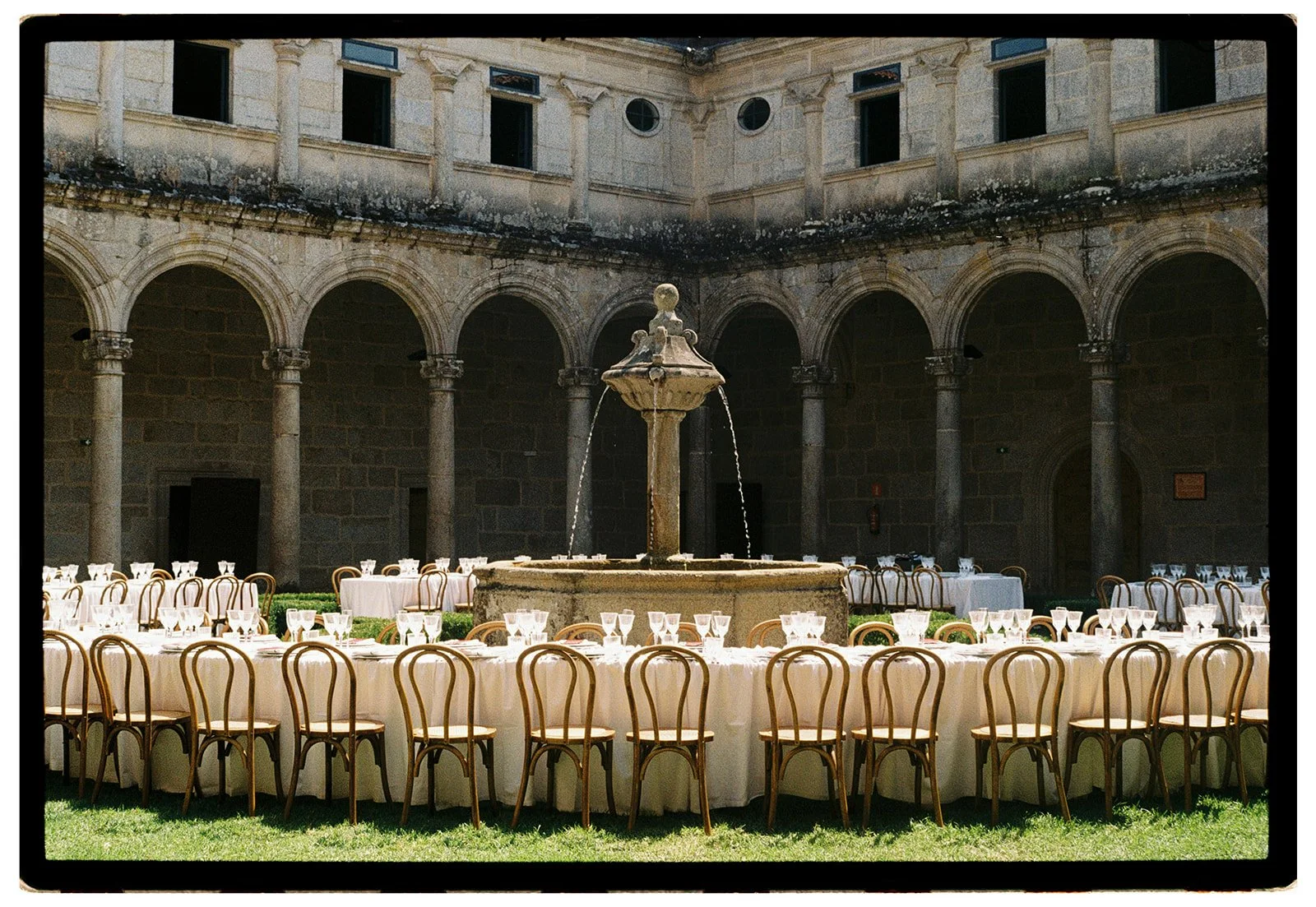 Outdoor courtyard set up for a formal event with round tables covered with white tablecloths, surrounded by wooden chairs, and a central stone fountain, with stone arches and windows on the building in the background.