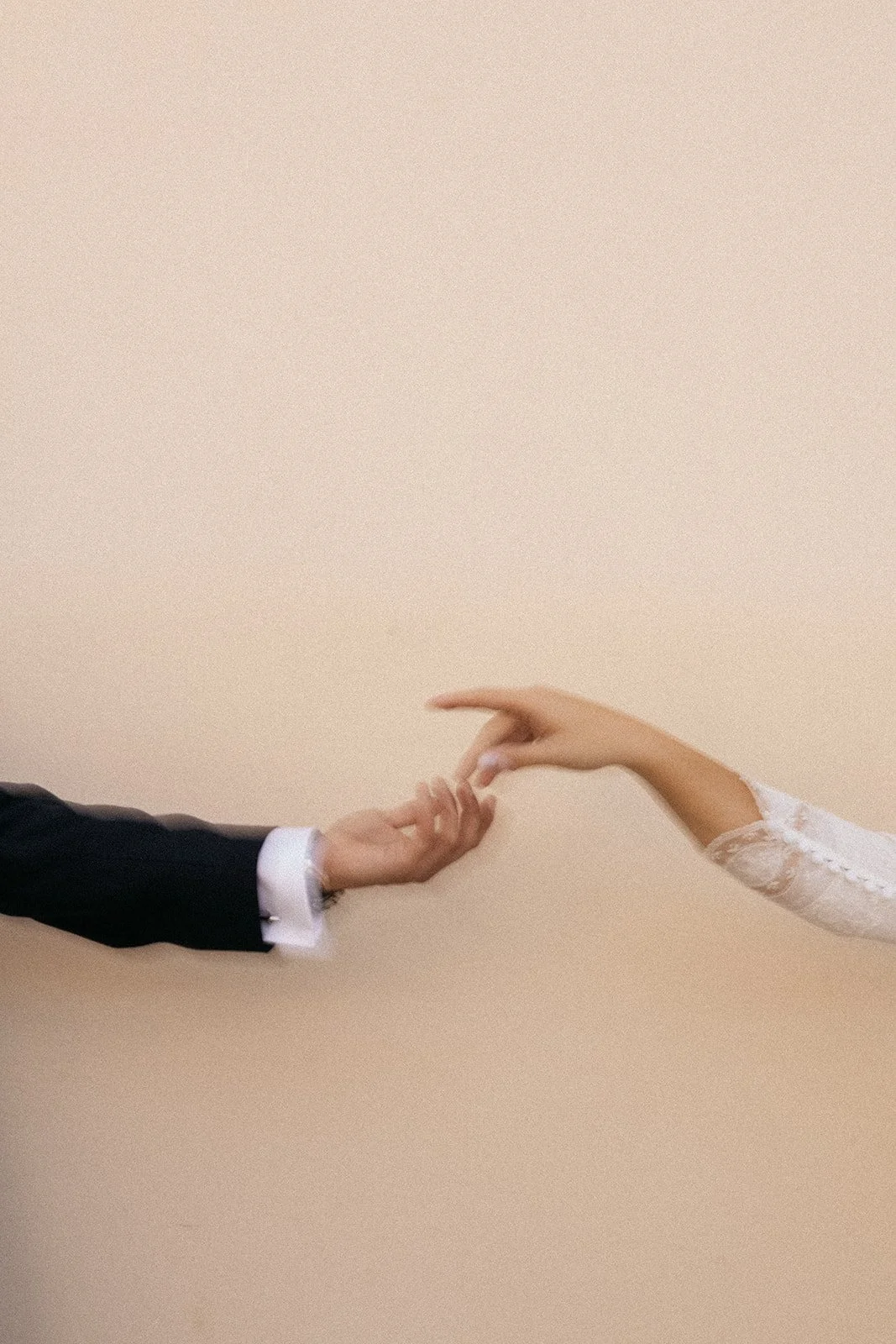 Close-up of a man and woman holding hands, with the man's hand in a suit and the woman's hand in a white lace dress, reaching toward each other.