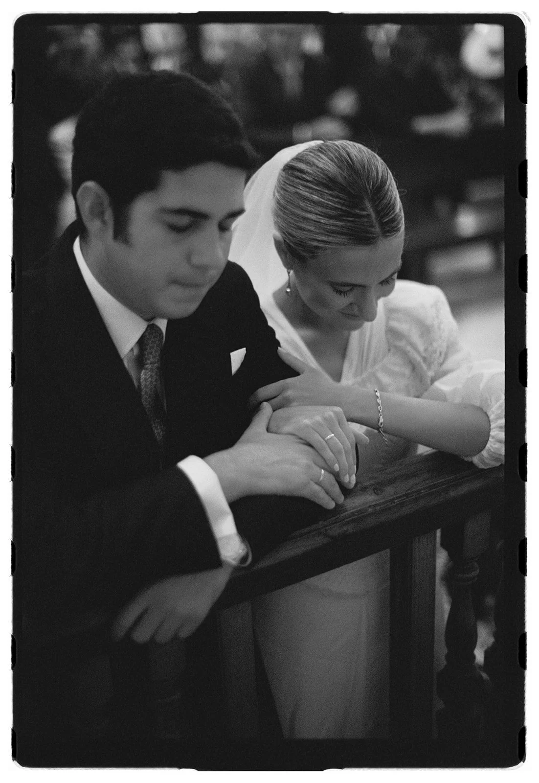 A black-and-white photo of a man and woman, likely at a wedding, holding hands and praying together with heads bowed, sitting at a wooden table in a church.